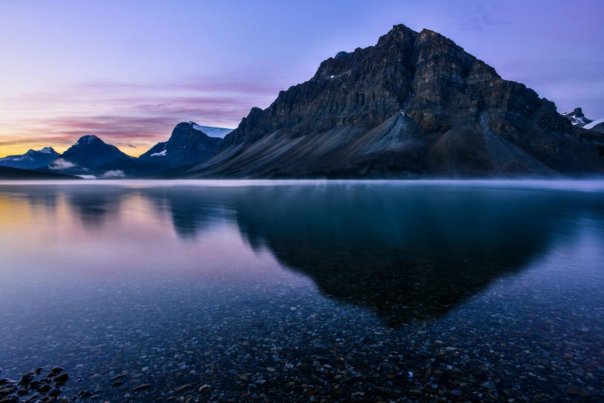 Rocky mountain range reflected in calm lake with clear water and colourful sky.