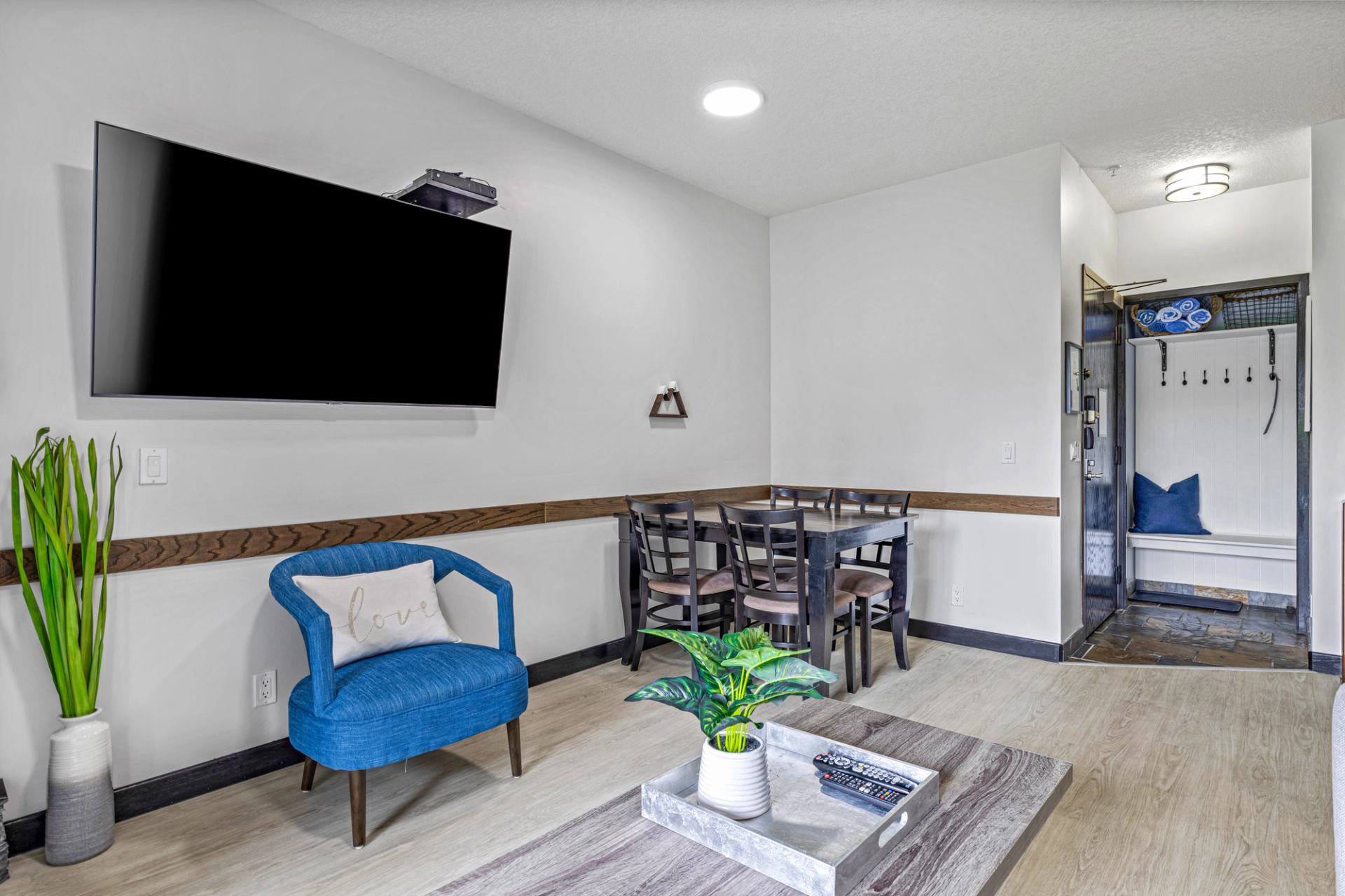 Living room with wall-mounted TV, blue chair, dining table and entryway bench.