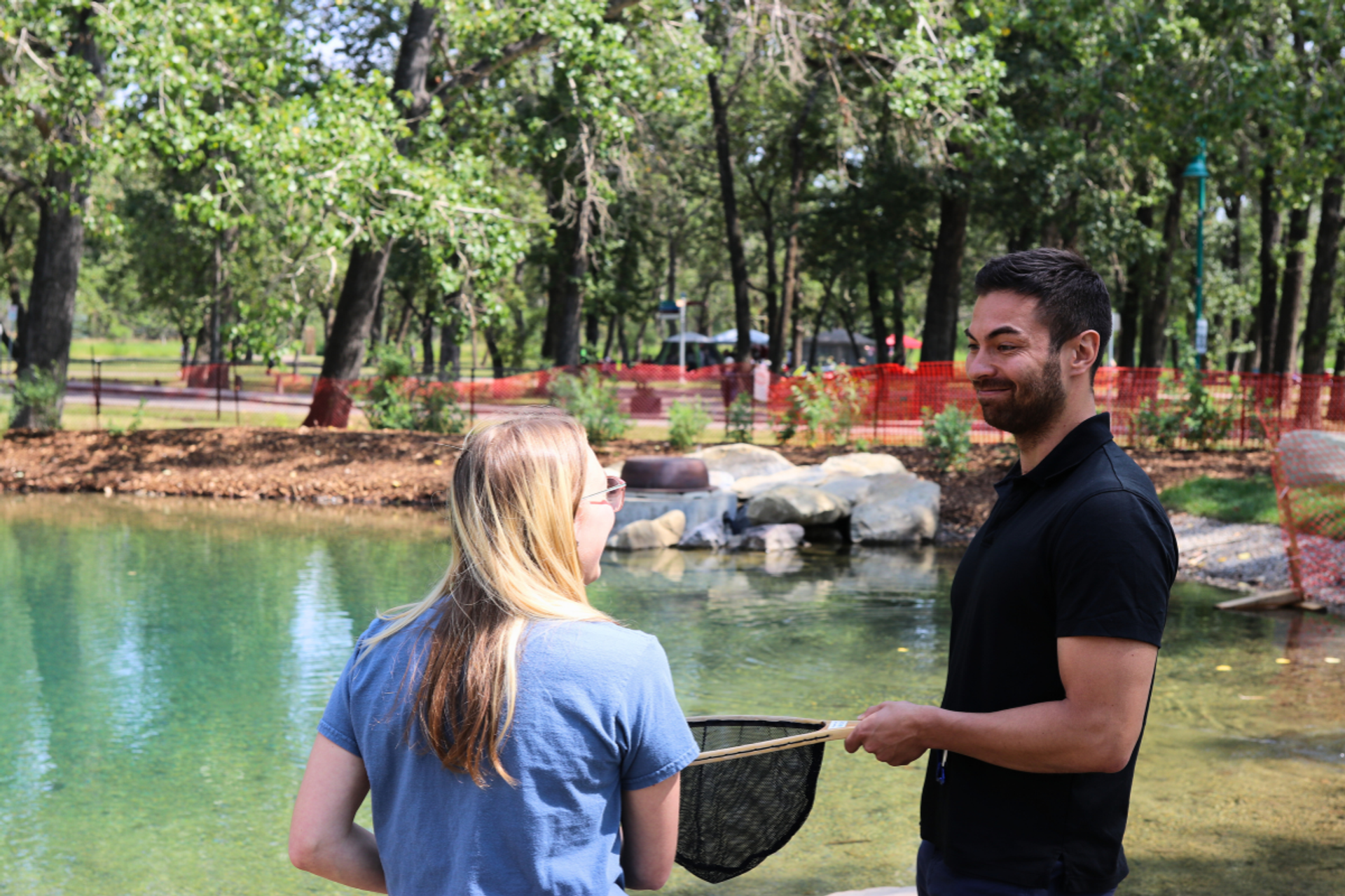 People fishing by an outdoor pond with a net in a wooded park setting.