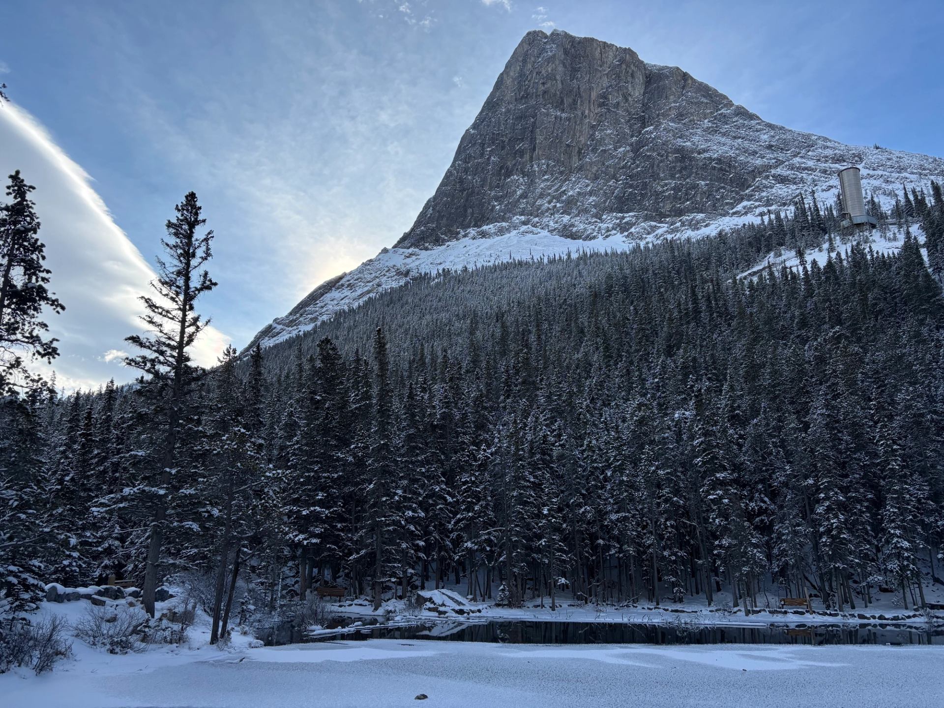 Snow-covered evergreen forest below steep rocky mountain beside frozen lake under blue sky.