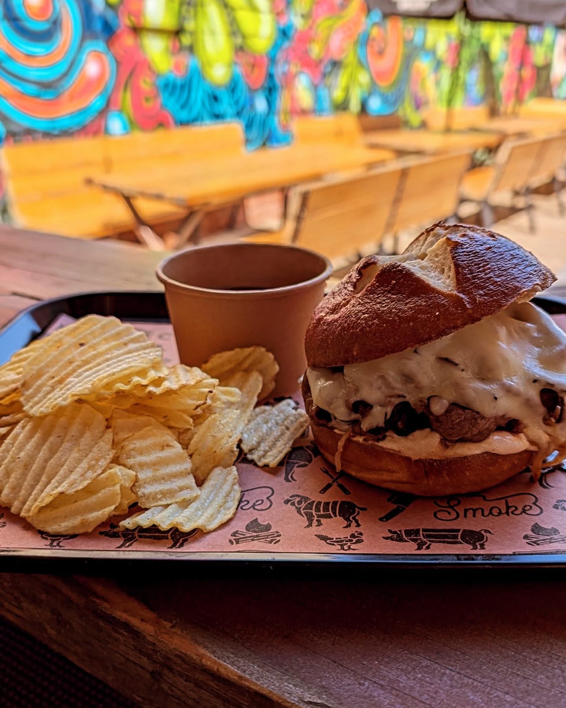 Burger with melted cheese and kettle chips sits on a tray before colourful patio mural artwork.