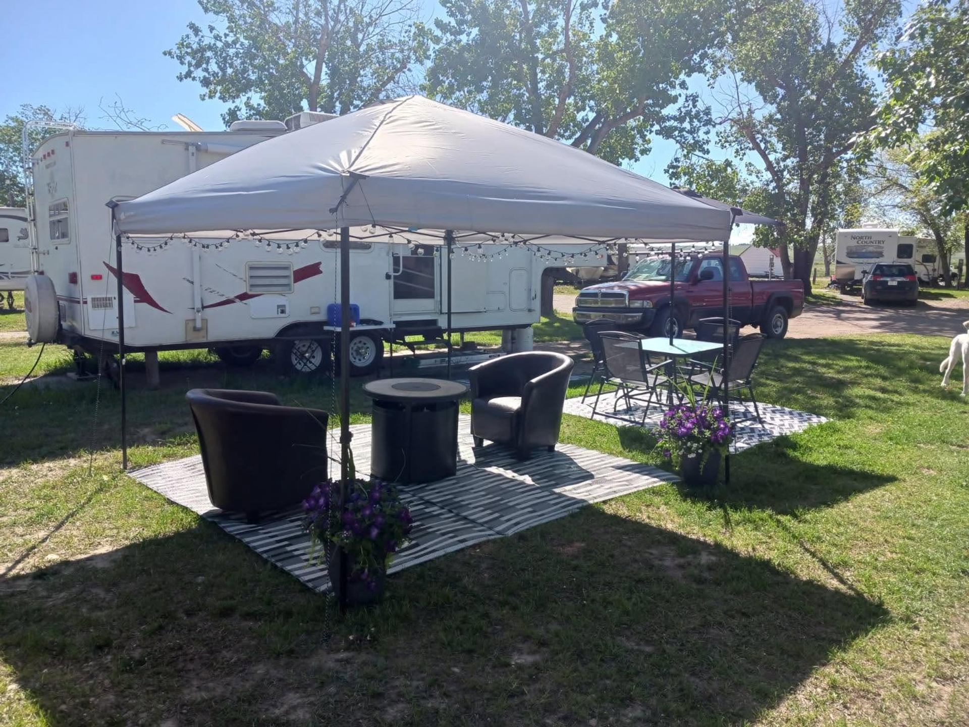 An RV campsite features a large canopy, outdoor lounge seating, flower planters, and shade trees.