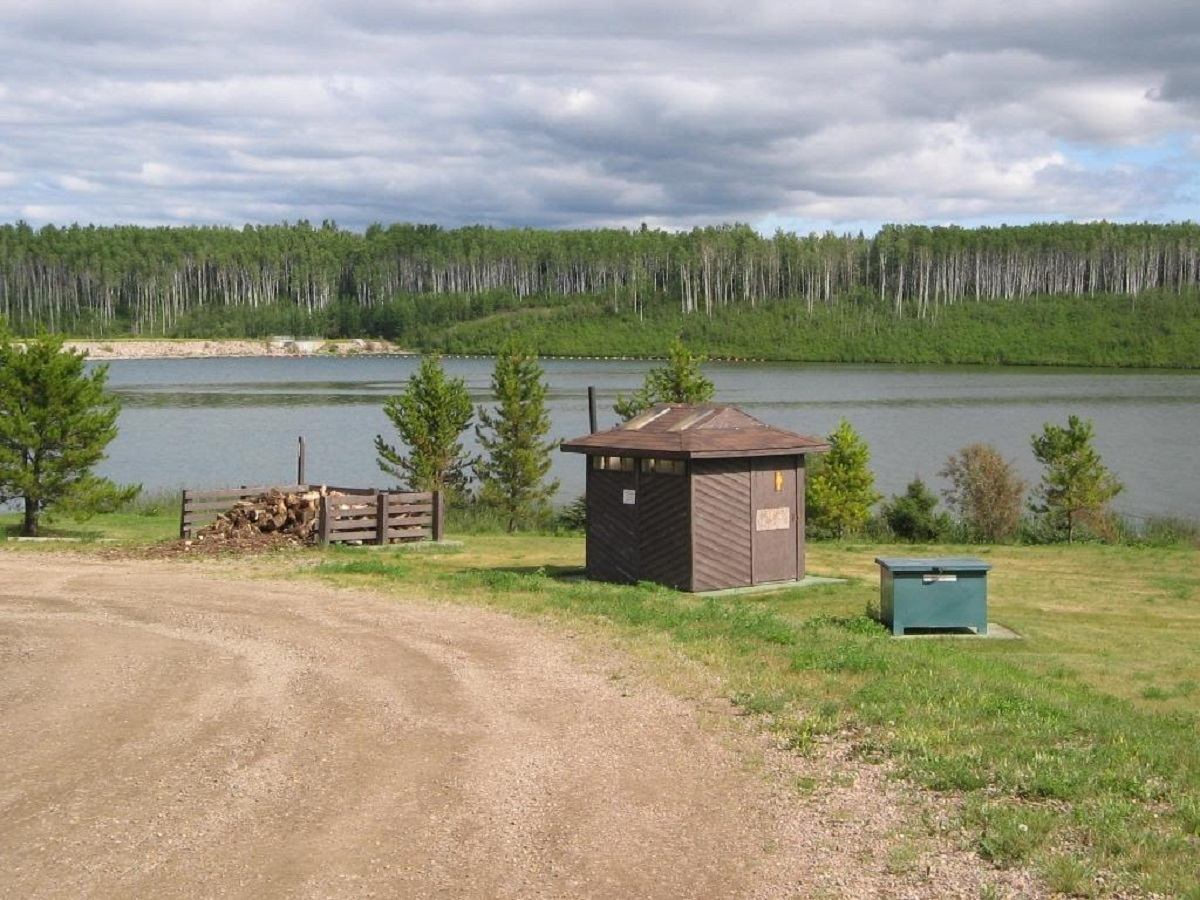 Small brown utility shed beside lake, with gravel road and forested shoreline behind.