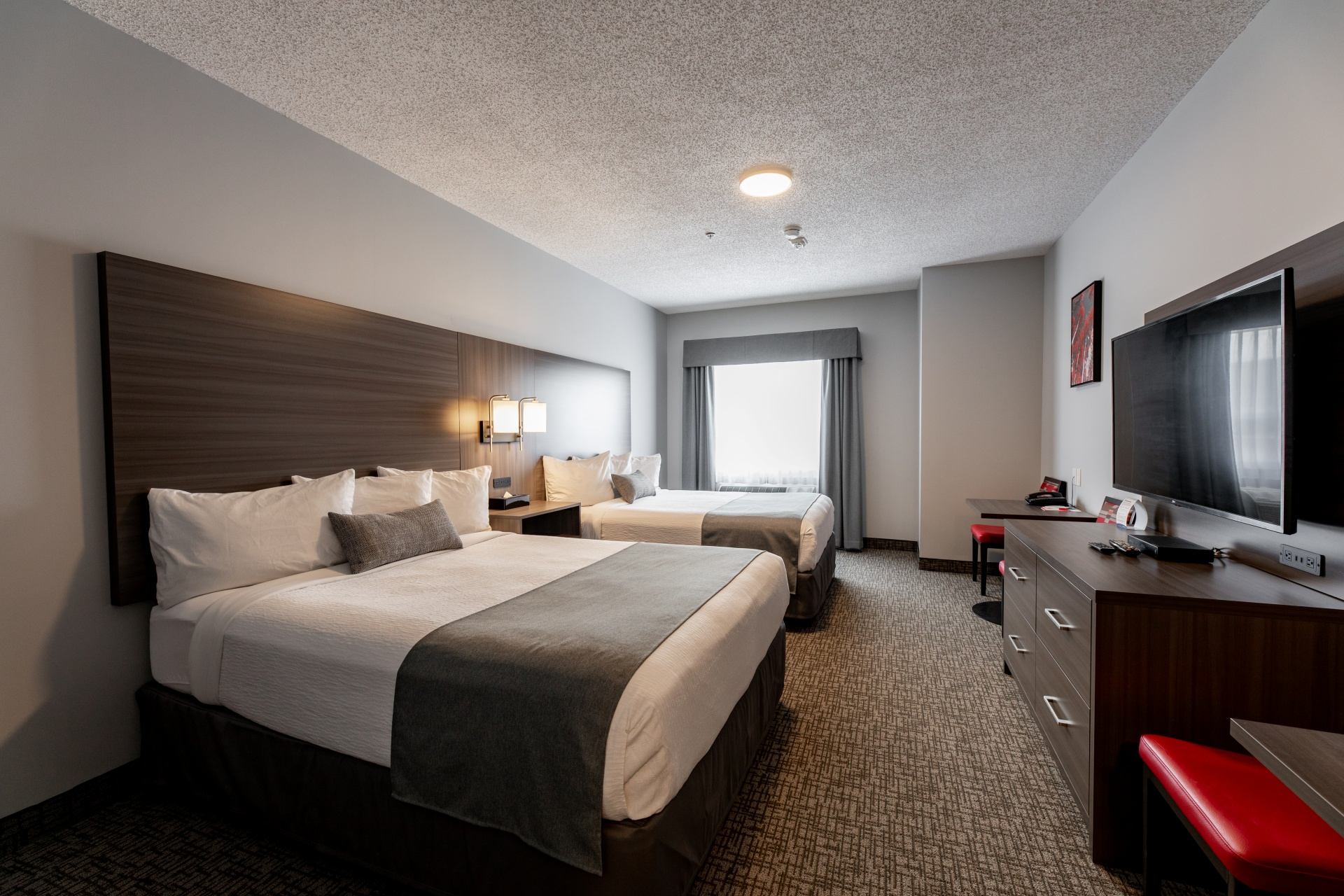 Hotel room with two beds, wall-mounted television, desk, dresser and red chairs.