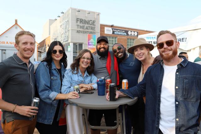 A group of 7 people gathered around a table in front of the Fringe Theatre Arts Barns: drinks in their hands and smiles on their faces!