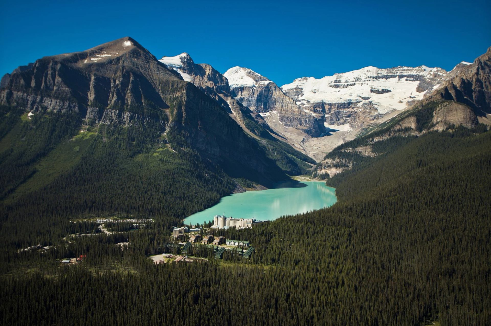 Turquoise mountain lake surrounded by dense evergreen forest and snow-capped rocky peaks.