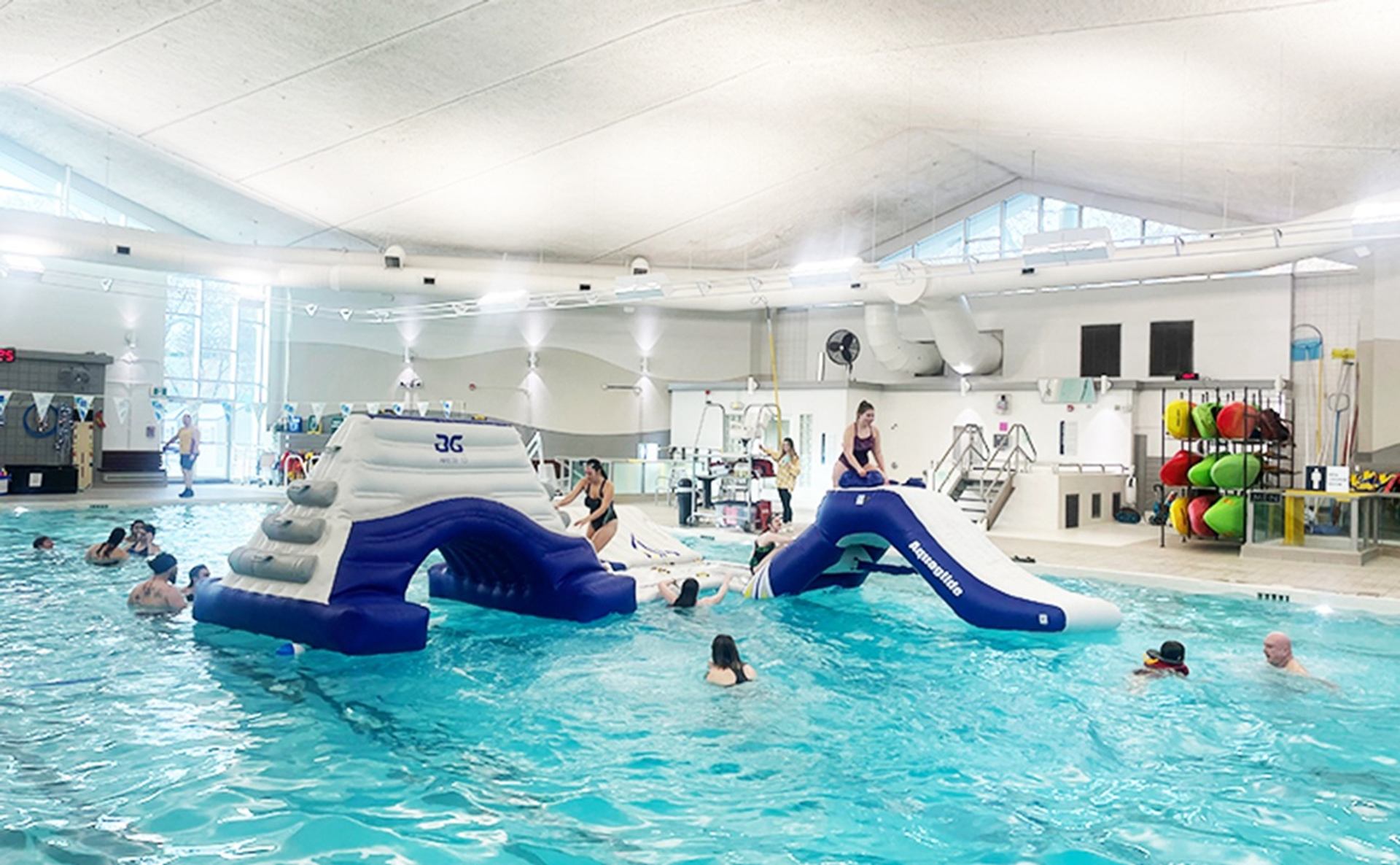 Indoor pool with inflatable climbing structure and slide, swimmers playing in turquoise water.