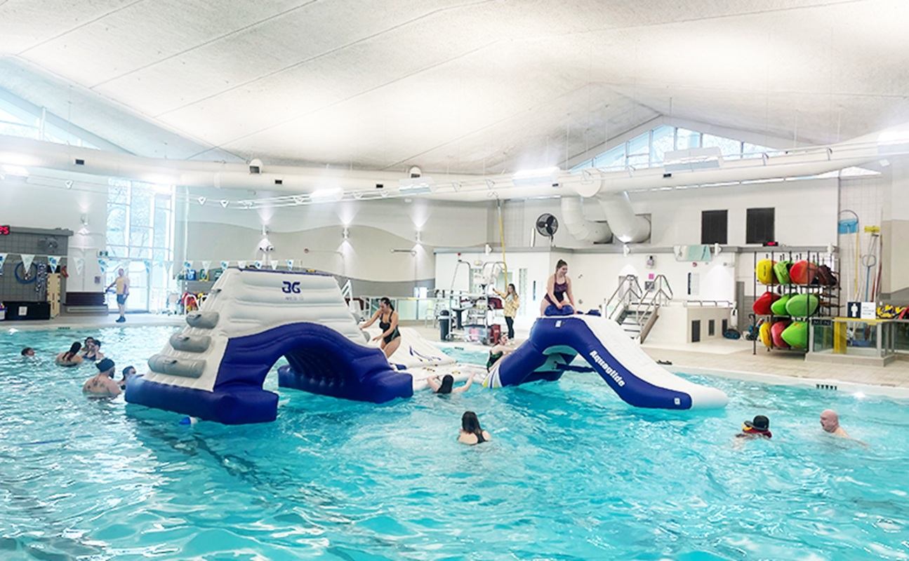 Indoor pool with inflatable climbing structure and slide, swimmers playing in turquoise water.