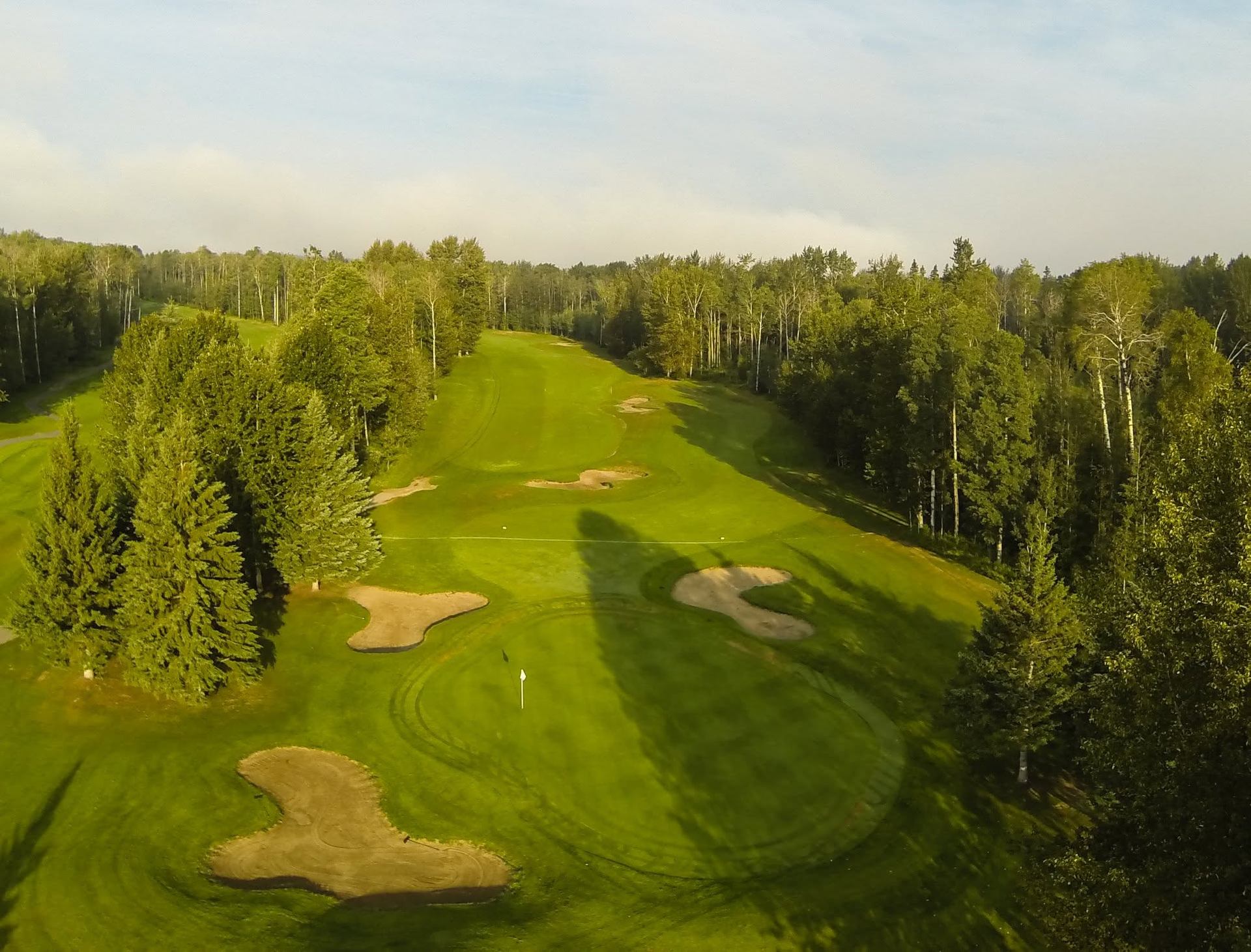 An aerial view showcases a tree-lined golf hole with manicured fairways, sand bunkers, and surrounding forest.