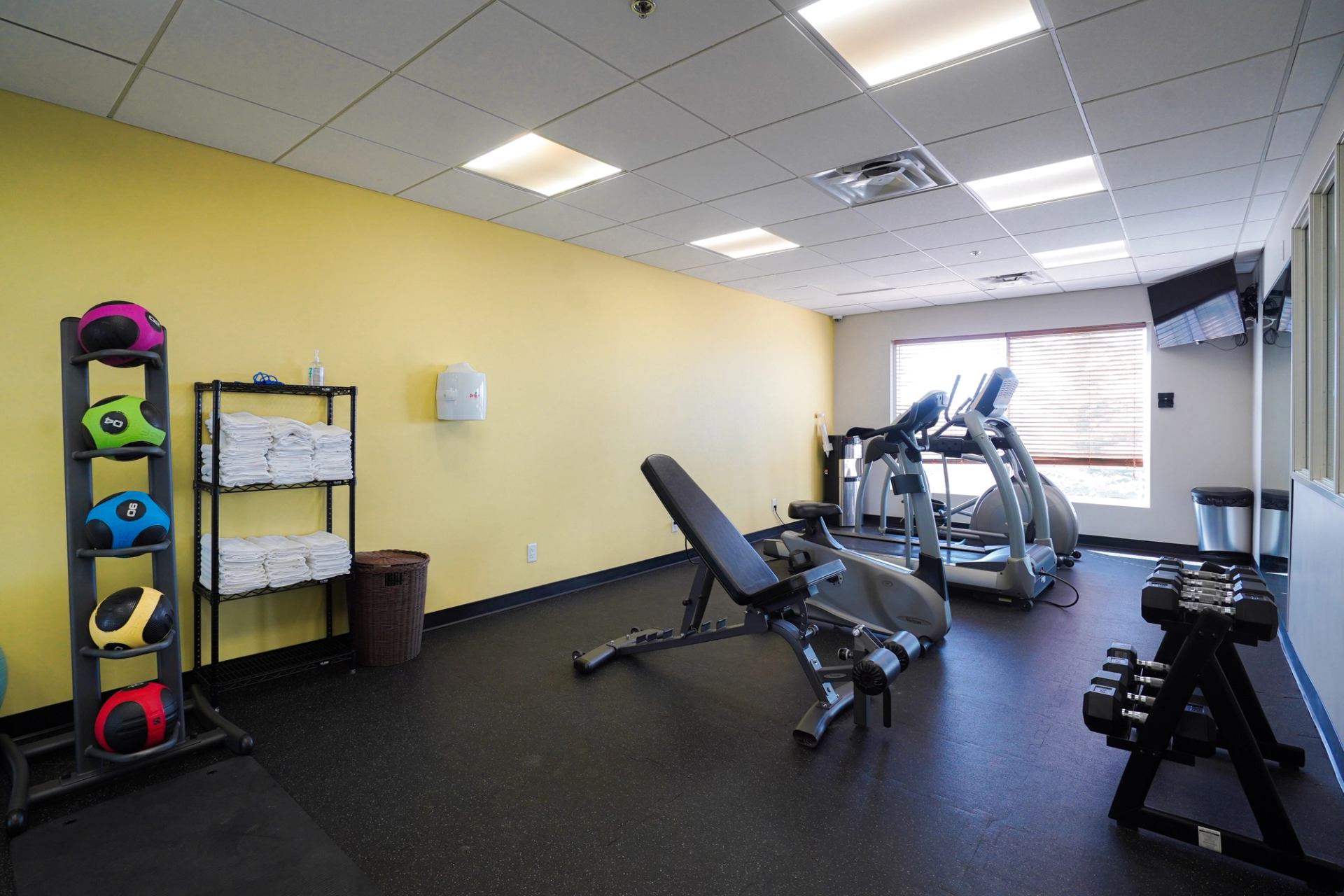 Fitness room with exercise machines, free weights, and medicine balls near a window.
