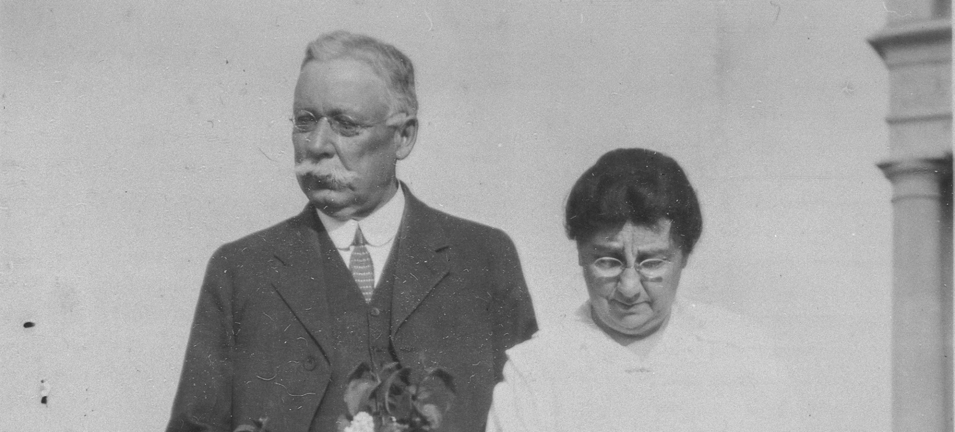 A black and white photo of an older man with a mustache and suit next to an older woman with glasses looking down.