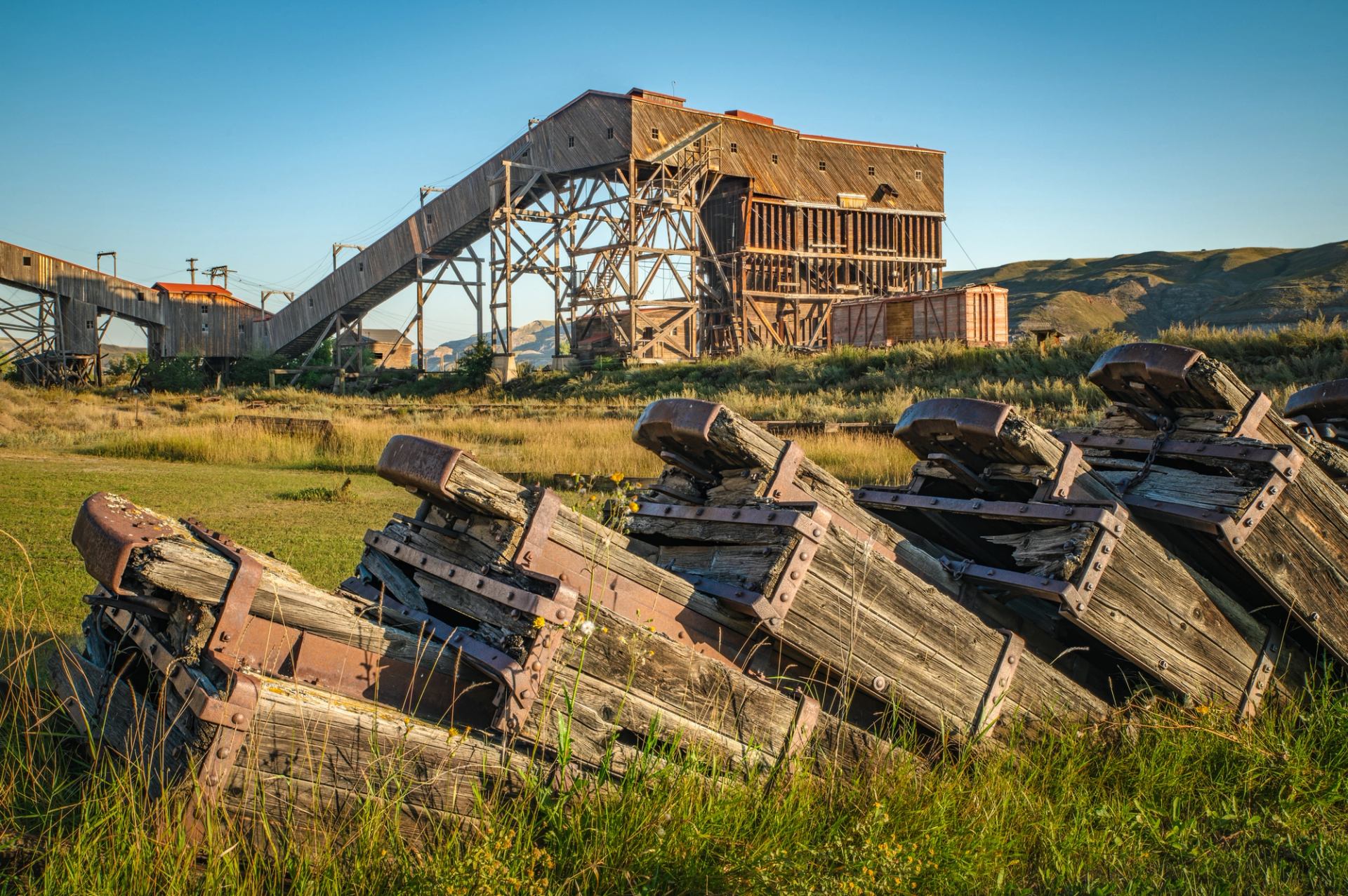 Abandoned wooden mining structure and rusted equipment in grassy field with distant hills.