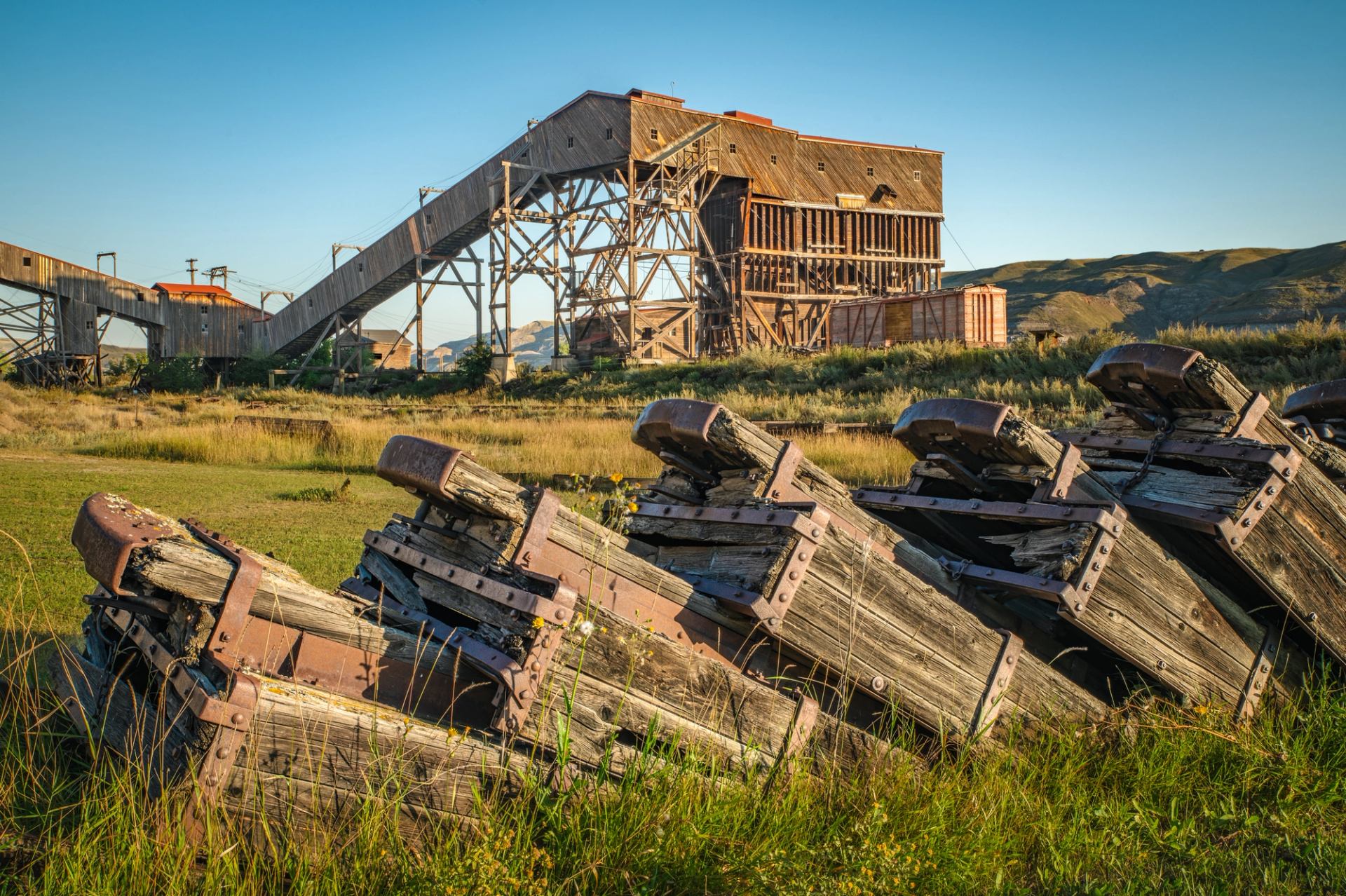 Abandoned wooden mining structure and rusted equipment in grassy field with distant hills.