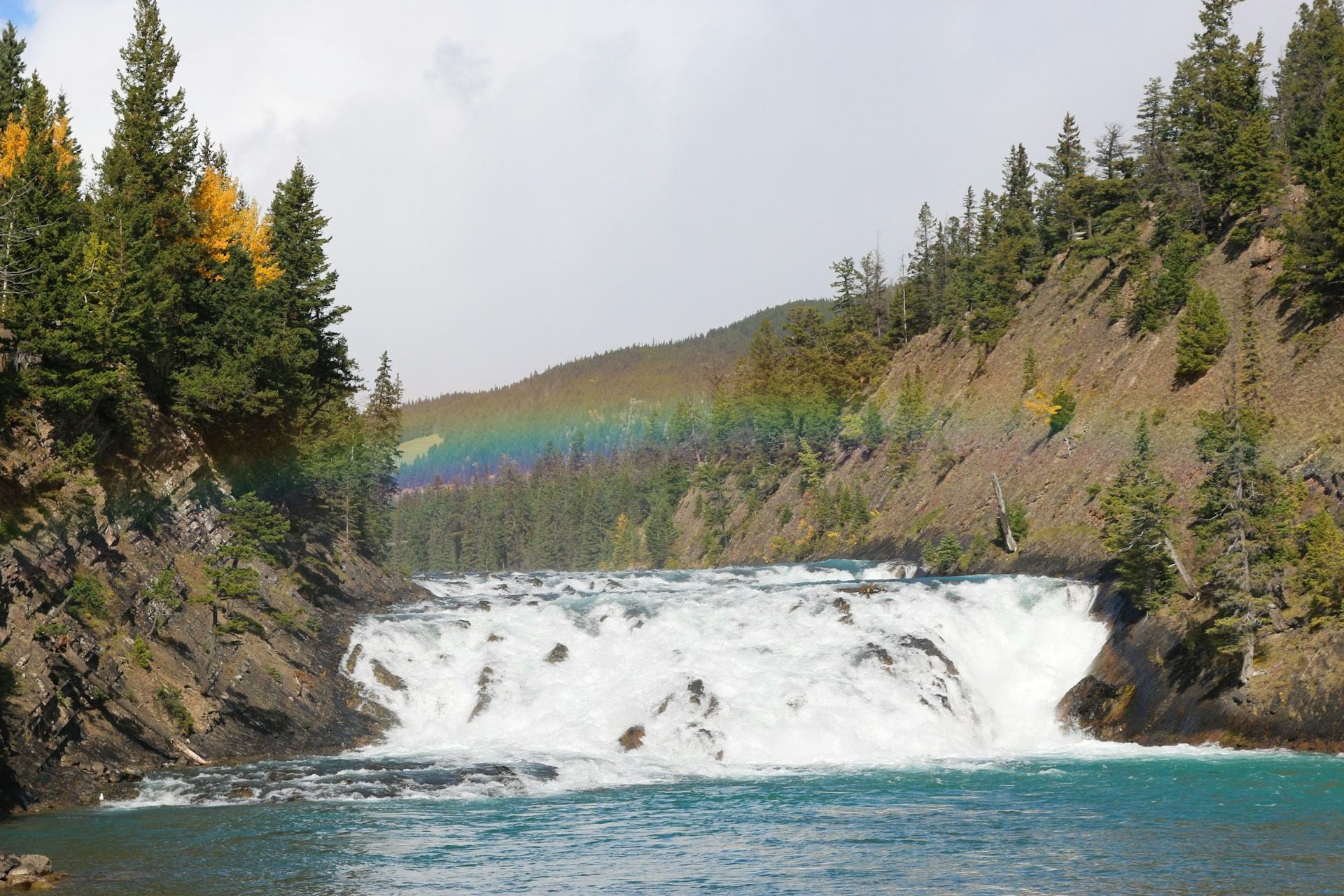 Wide waterfall flowing between forested cliffs with a faint rainbow arching above the trees.