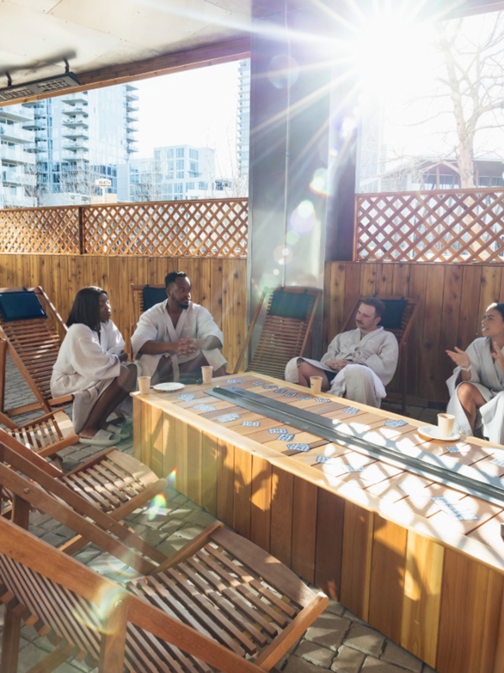 People in robes sit on wooden lounge chairs around outdoor table with playing cards.