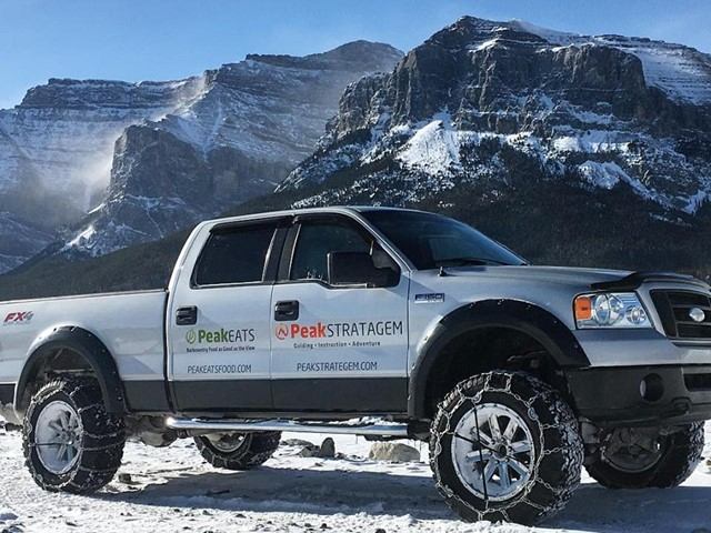 A silver pickup with branded logos and tire chains sits in snowy terrain, mountains towering under a clear blue sky.