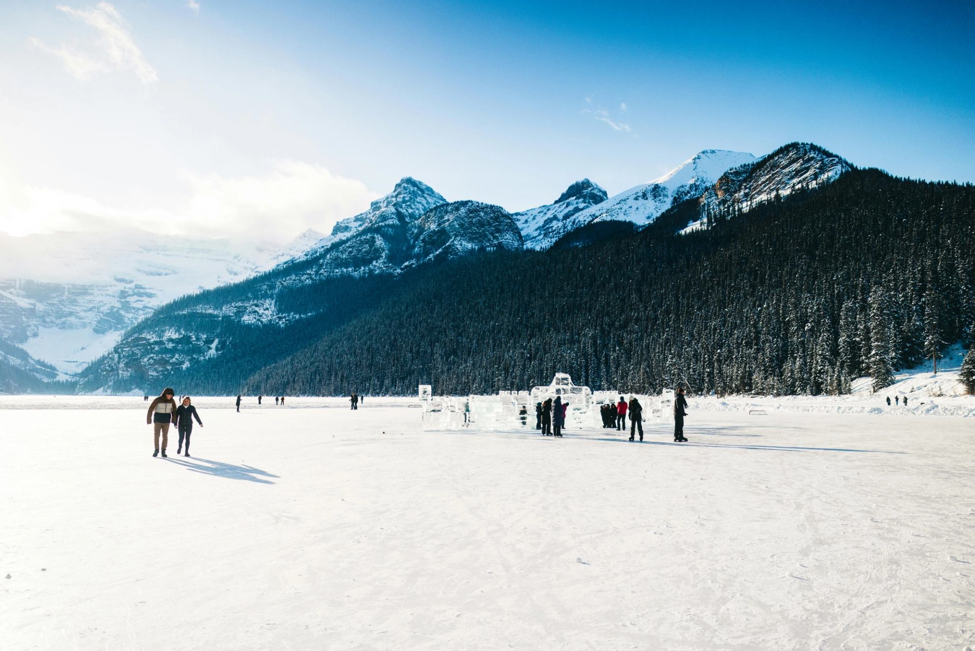 A winter scene of people walking on a vast frozen lake with ice sculptures, framed by snow-covered mountains and pine forests.