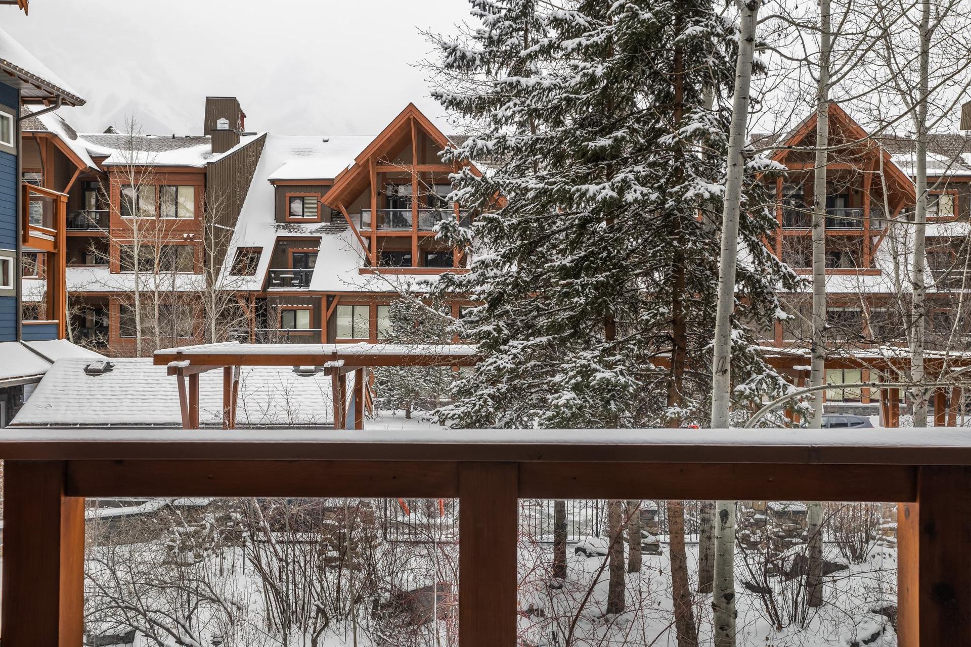 Snow-covered lodge-style buildings and trees viewed from a wooden balcony railing.