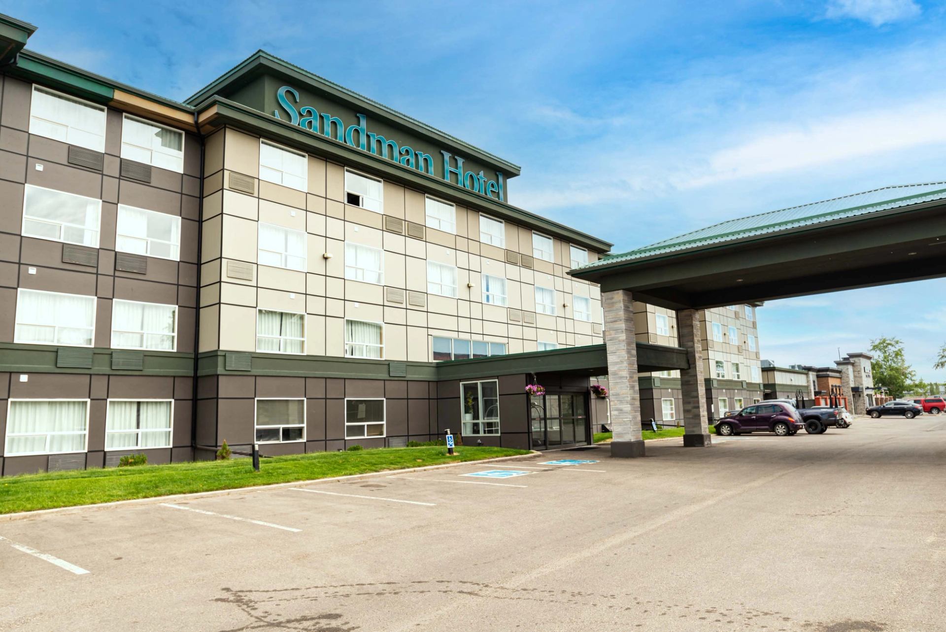 Sandman Hotel exterior with covered entrance, parking lot and several parked vehicles.