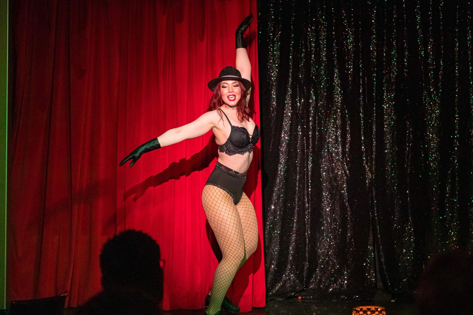 Burlesque performer in black costume poses on stage before red and black curtains.