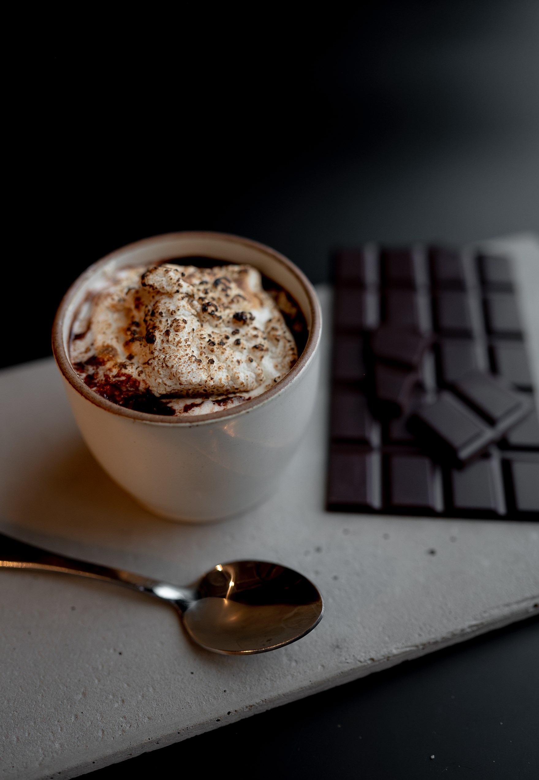 Cup of hot chocolate with foam, spoon on a tray, blurred chocolate blocks in background.