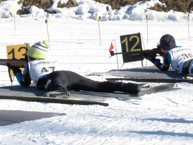 Biathletes lying on snow aim rifles at numbered targets on an outdoor shooting range.