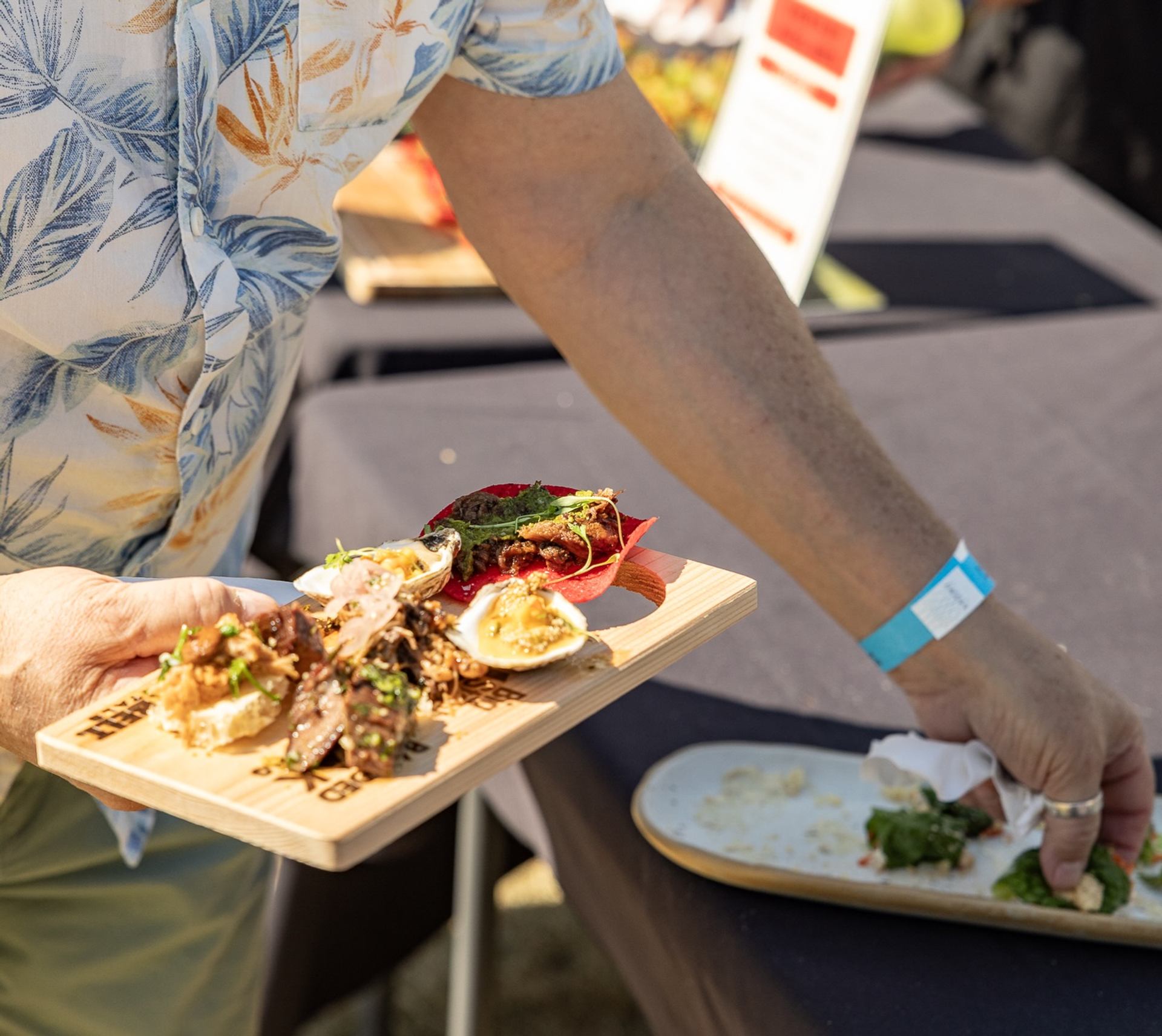 Person in patterned shirt carries wooden board with assorted appetizers at outdoor event.