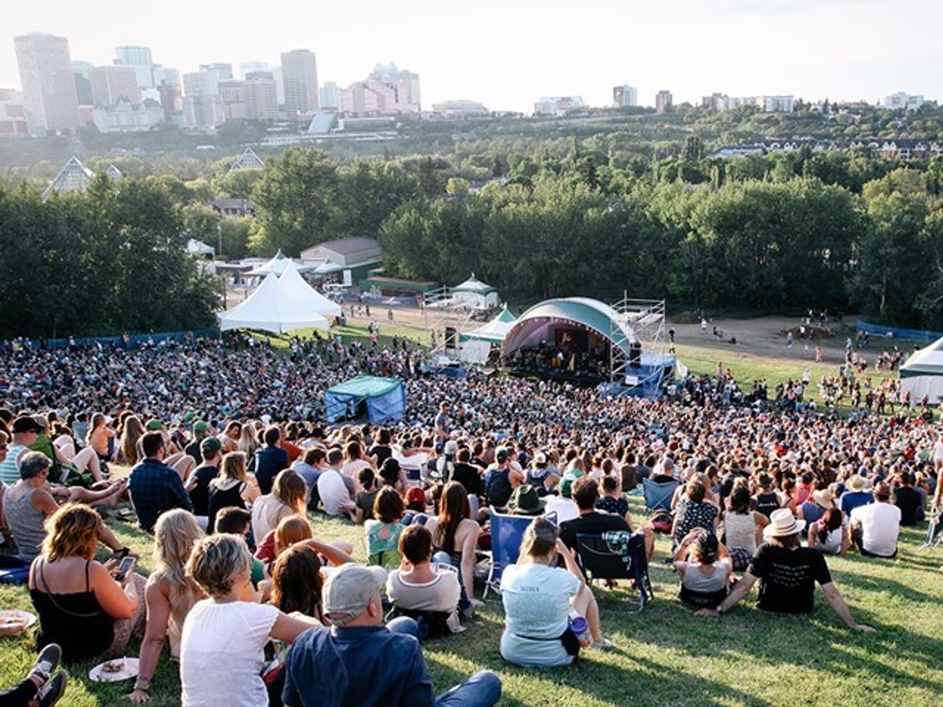 People seated on grass near a stage with tents and Edmonton skyline in background