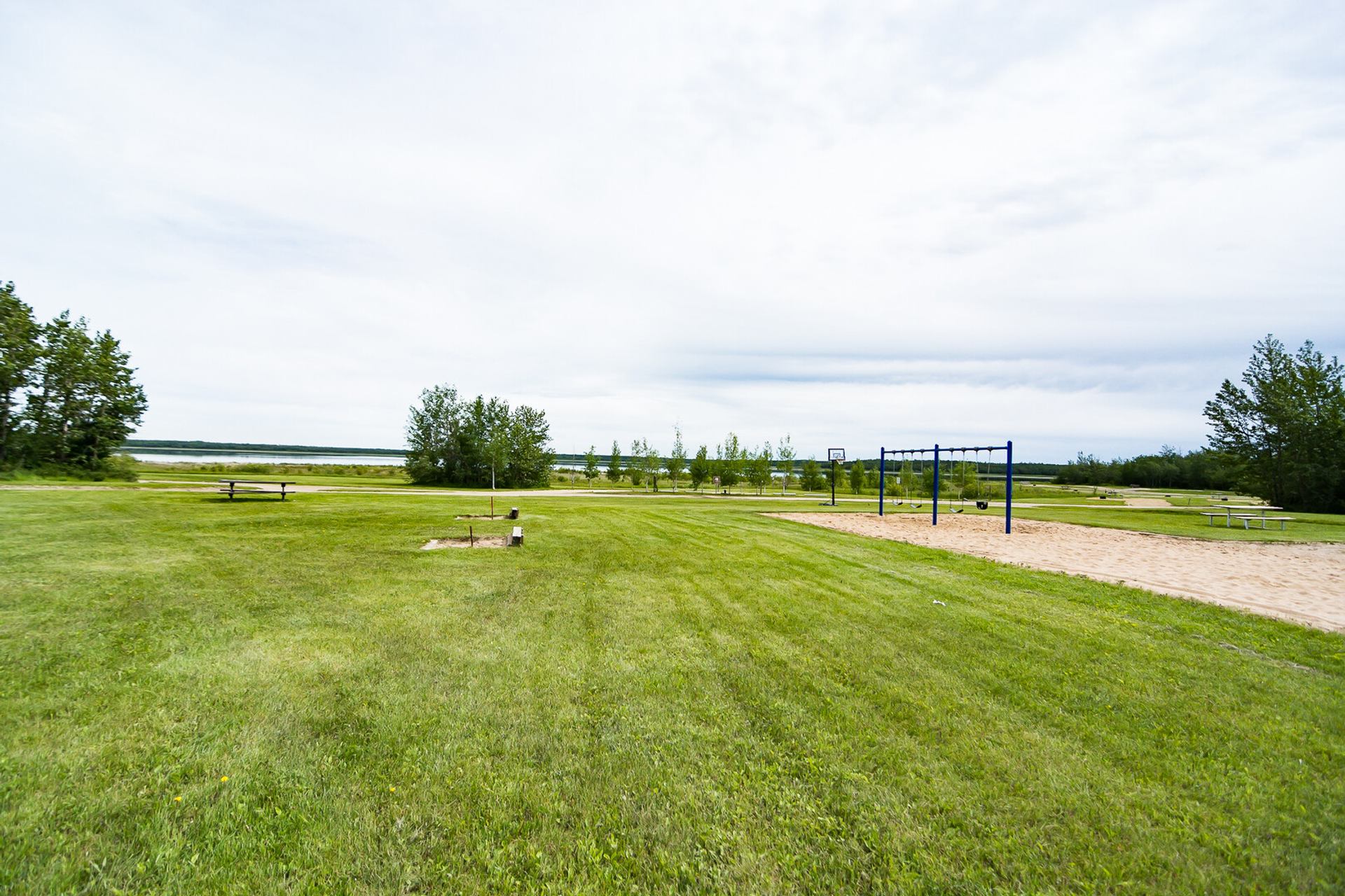 A grassy park with a swing set, picnic table, and horseshoe pits, overlooking a lake under a cloudy sky.