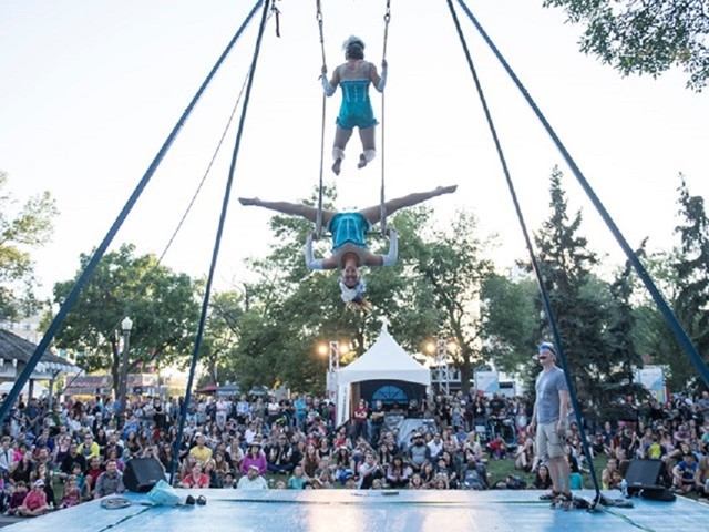 Aerial performers in blue swing on trapeze against clear sky; crowd watches below amid trees and festival tents, lively atmosphere.
