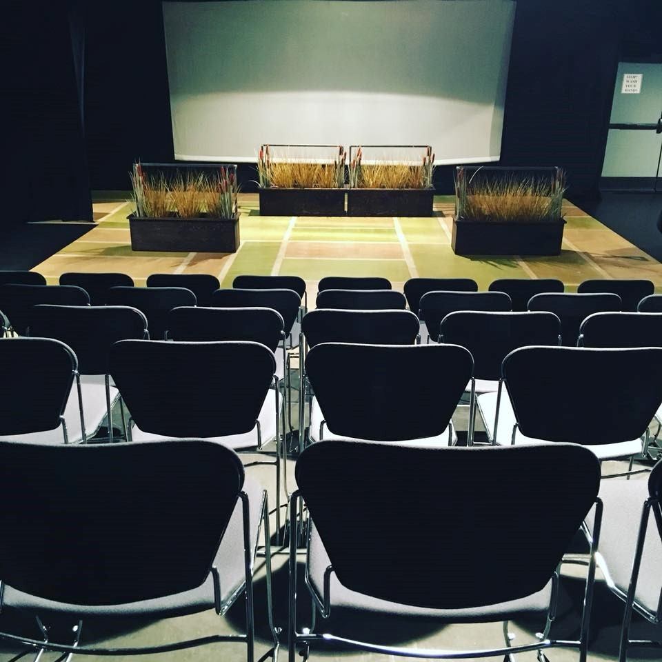 Rows of empty chairs facing a stage with planters and a large projection screen.
