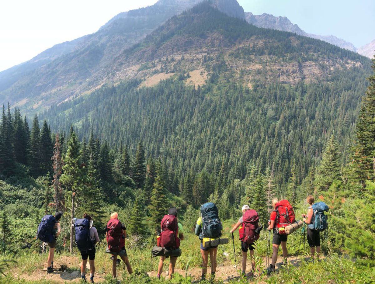 A group of backpackers walking along a mountain trail above a dense evergreen valley.