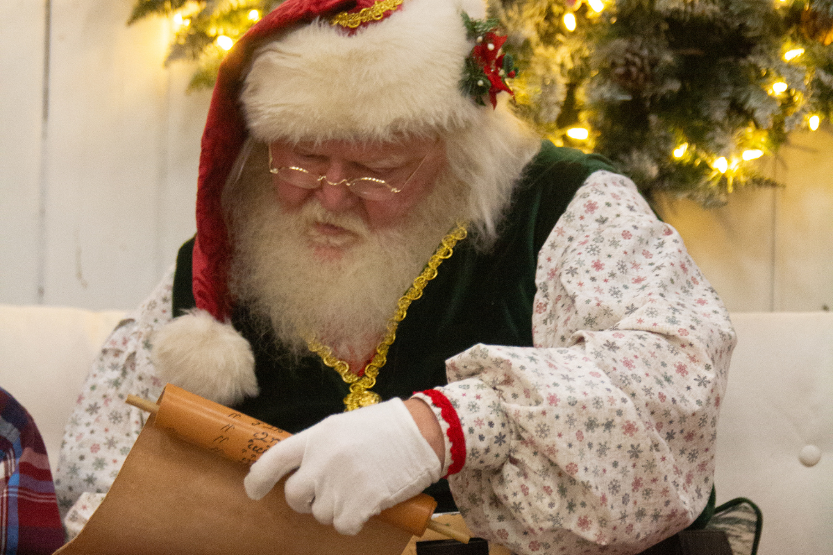 Santa Claus in festive attire reads a scroll amid Christmas lights and decorations.