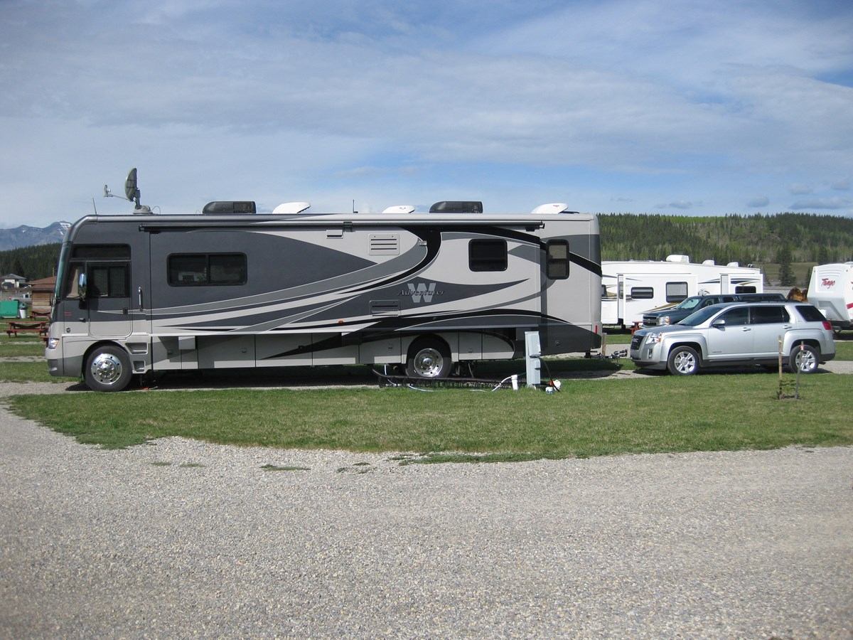 Large motorhome and SUV are parked at a serviced RV campsite with grassy grounds and nearby RVs.