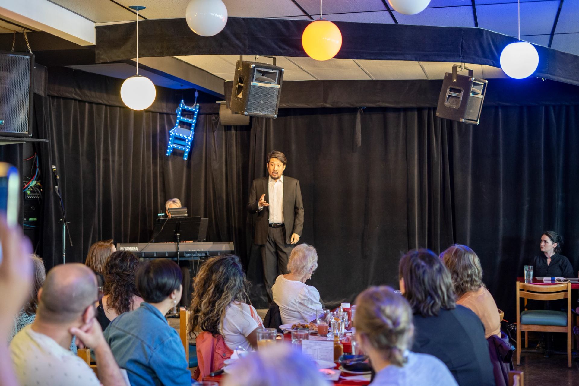 A man in a suit performs opera on stage at the Blue Chair, with a pianist accompanying him on keyboard.