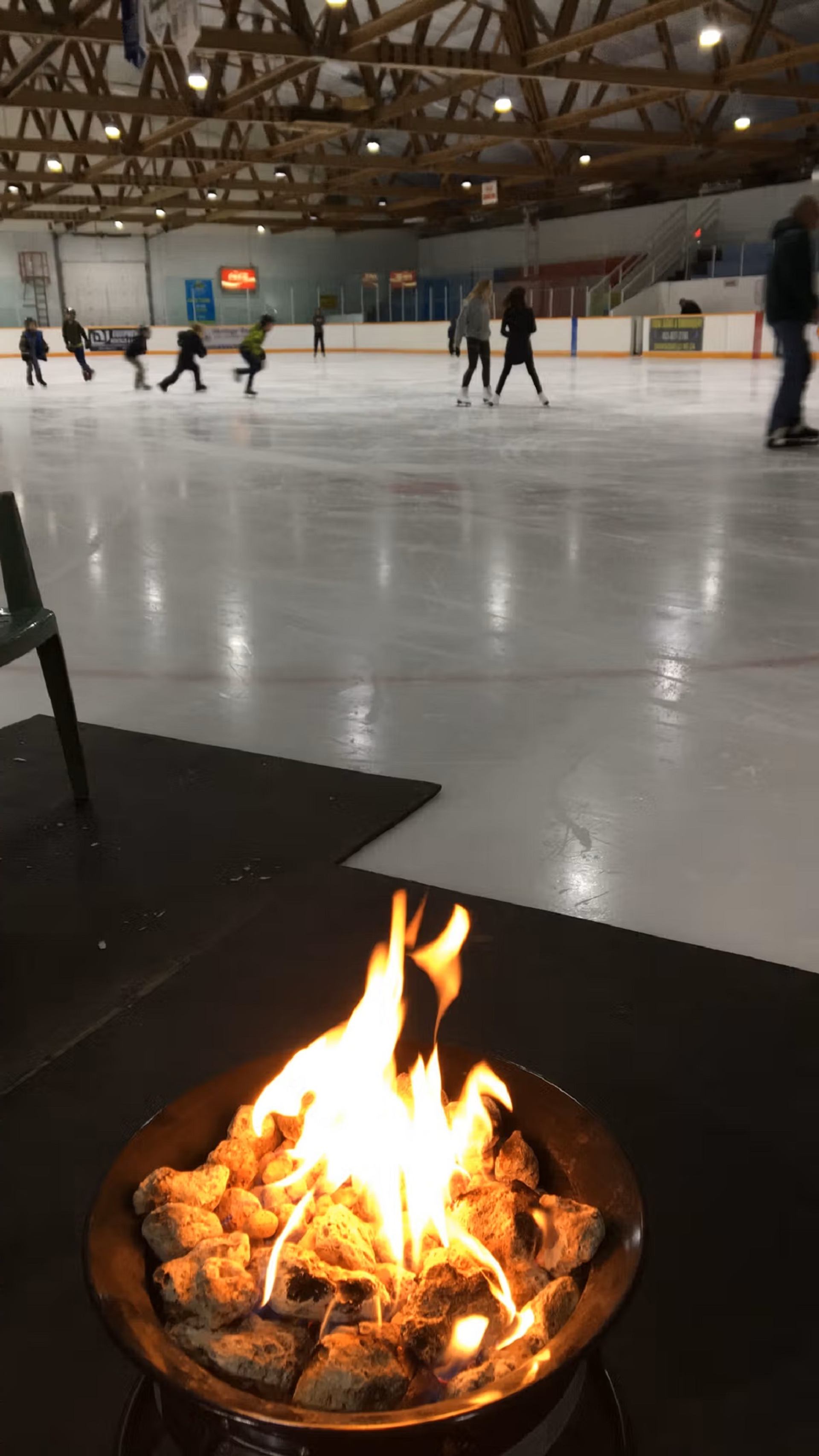 A fire pit with flames in the foreground, with an ice rink and skaters in the background.