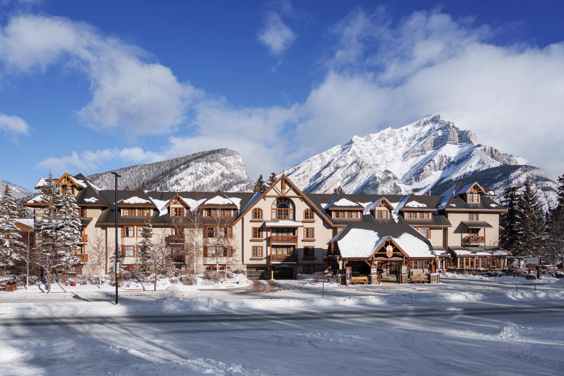 Hotel with snow on the roof, with snow capped mountains behind and a cloudy blue sky.