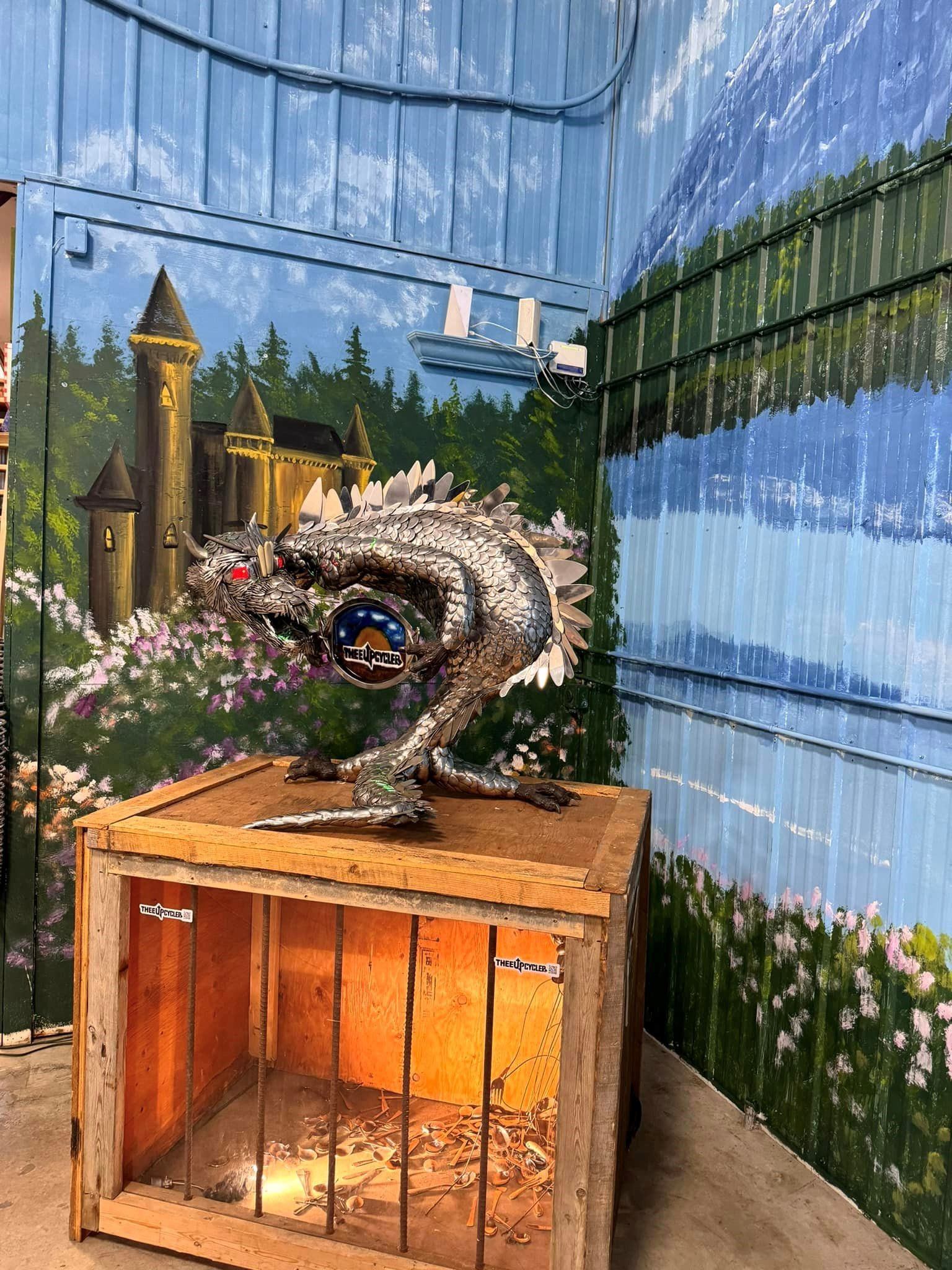 Metal dragon sculpture on wooden display, against mural of castle and forest.