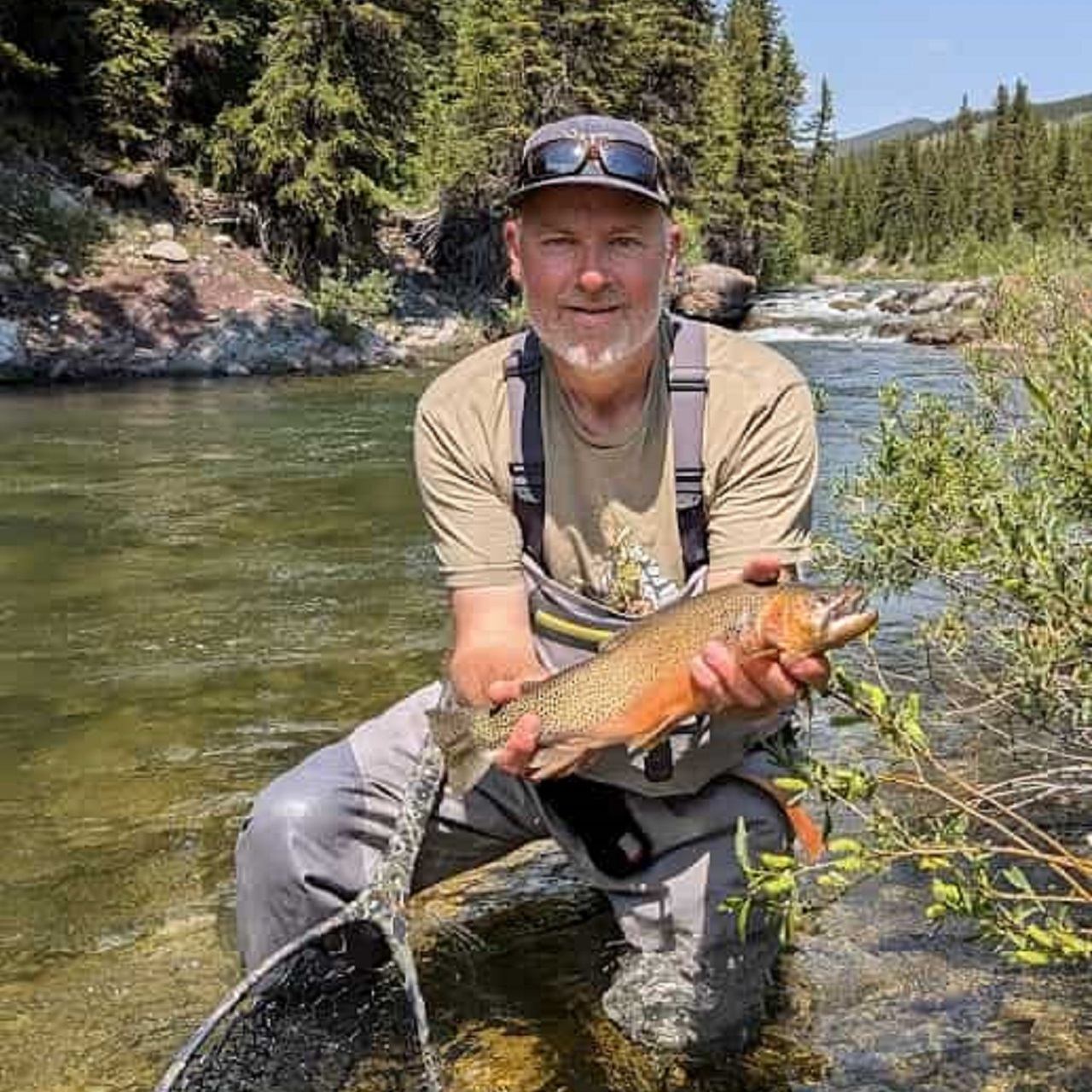 A man in fishing waders kneels in a river, holding a colorful trout.