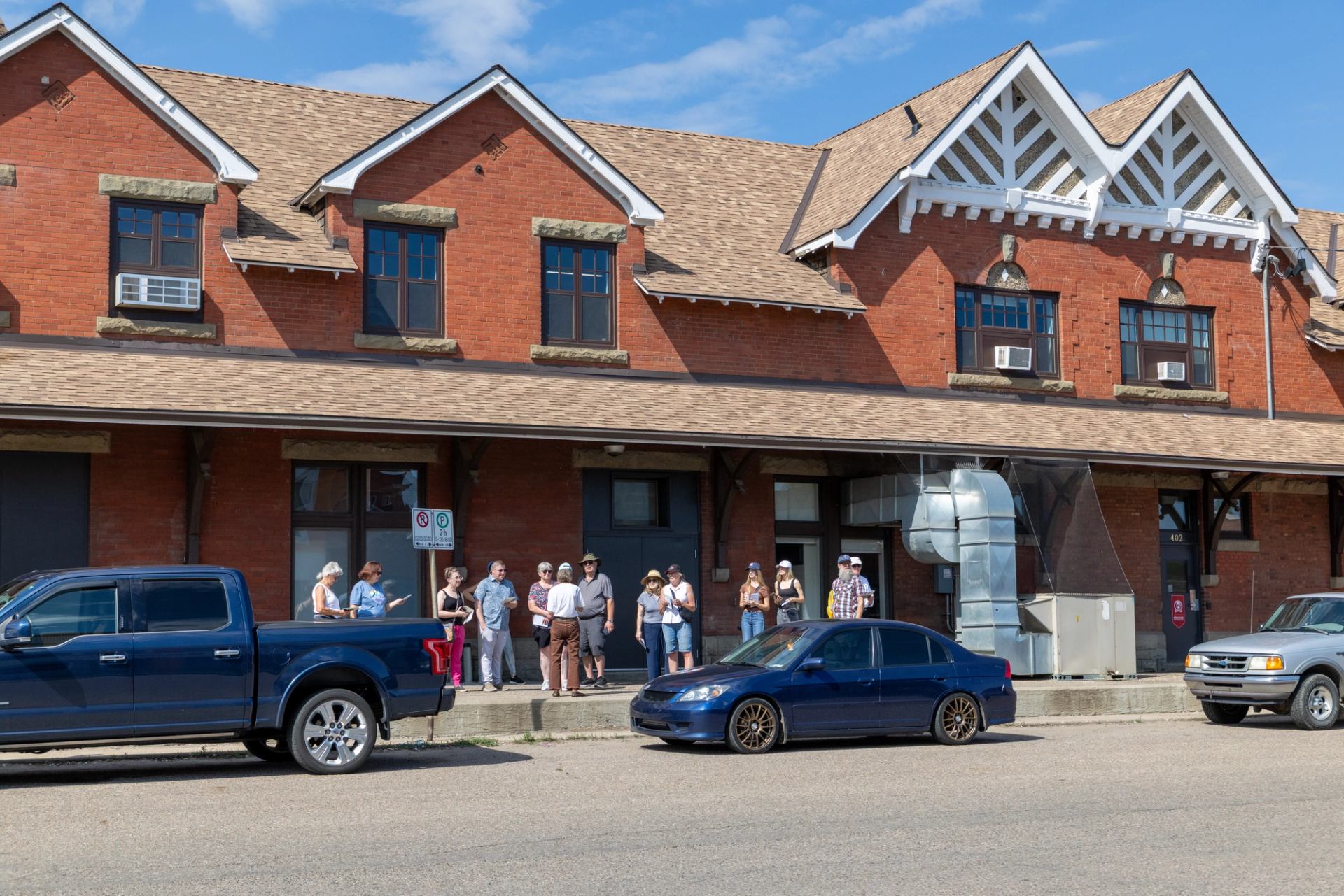 Group of people standing outside brick building with parked vehicles along street.