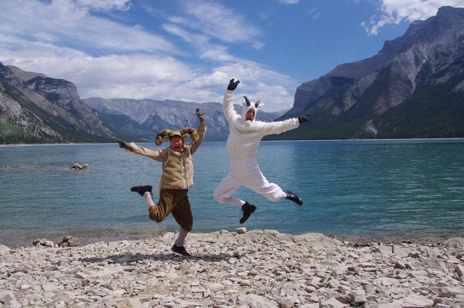 Two costumed mascots jumping on a rocky lakeshore with mountains.