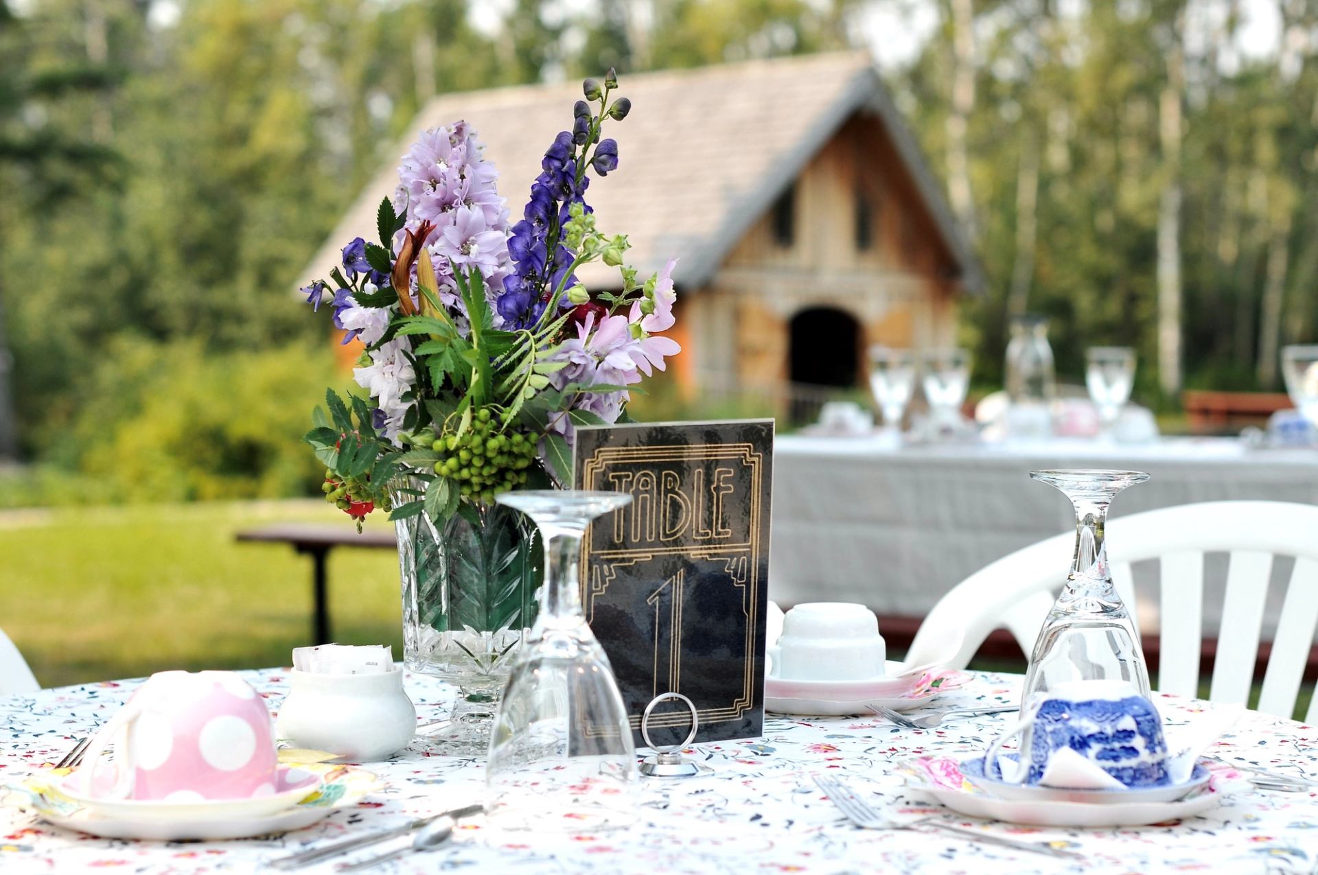 Table with floral centerpiece, glassware, and rustic building in background