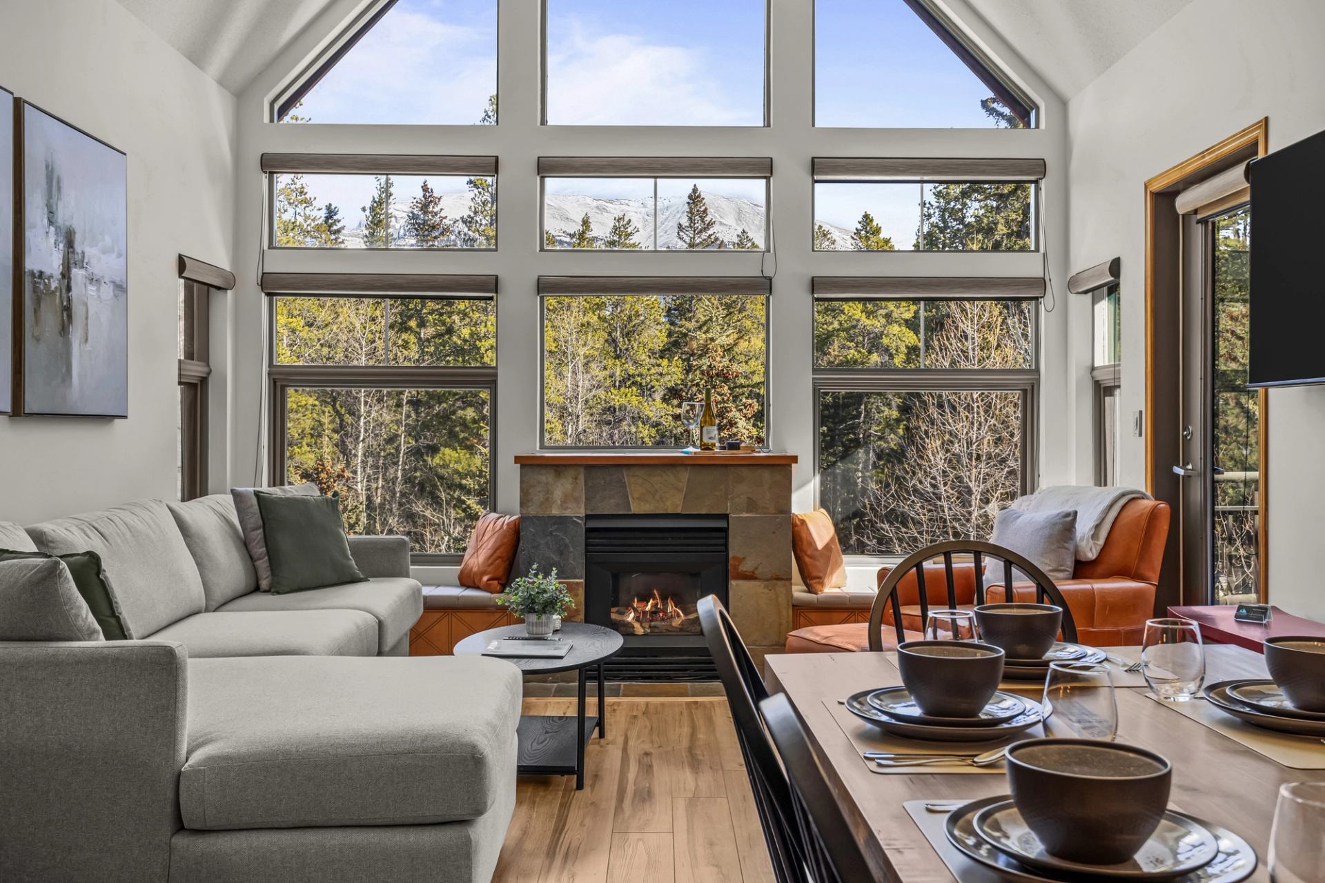 A spacious living area features a large sectional sofa, stone fireplace, dining table, and floor-to-ceiling windows overlooking a forest and distant mountain peaks.