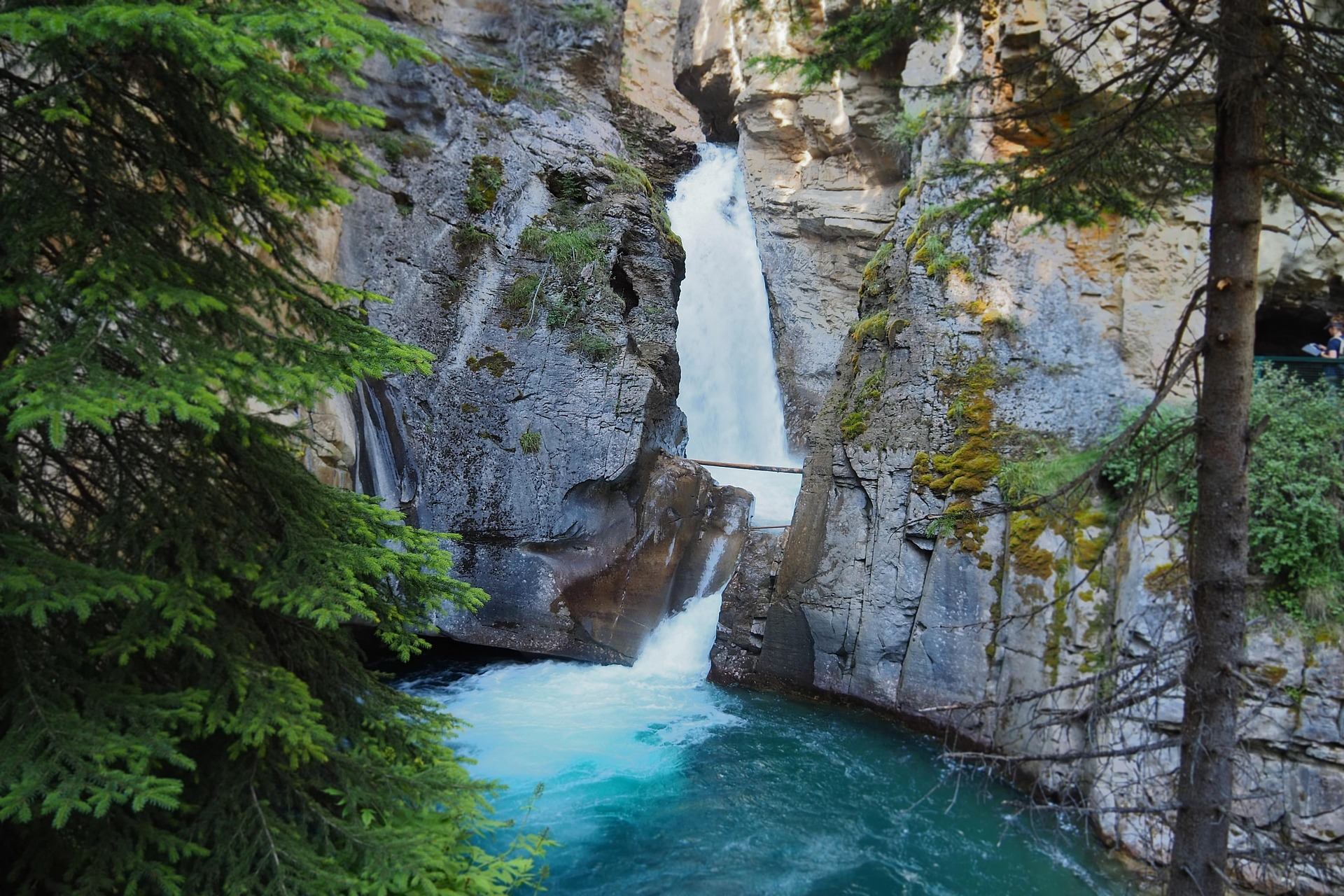 Waterfall plunging between rocky cliffs into turquoise pool, framed by evergreen trees and moss.
