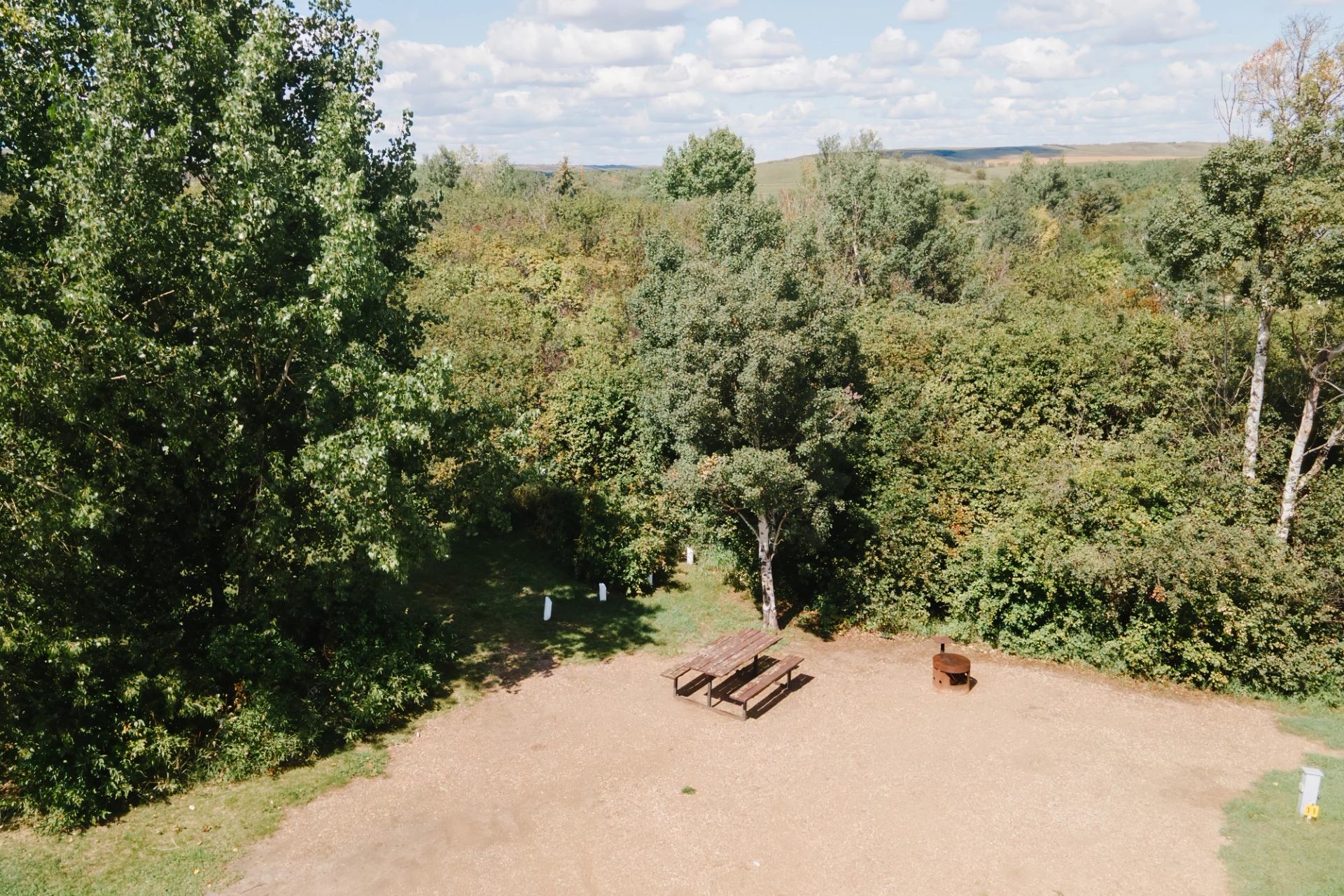 Gravel campsite with wooden picnic table and fire ring beside dense green trees.