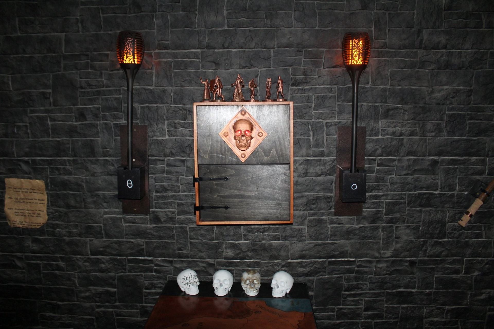 Stone wall with glowing skull panel, torches, figurines, and skulls on wooden table.