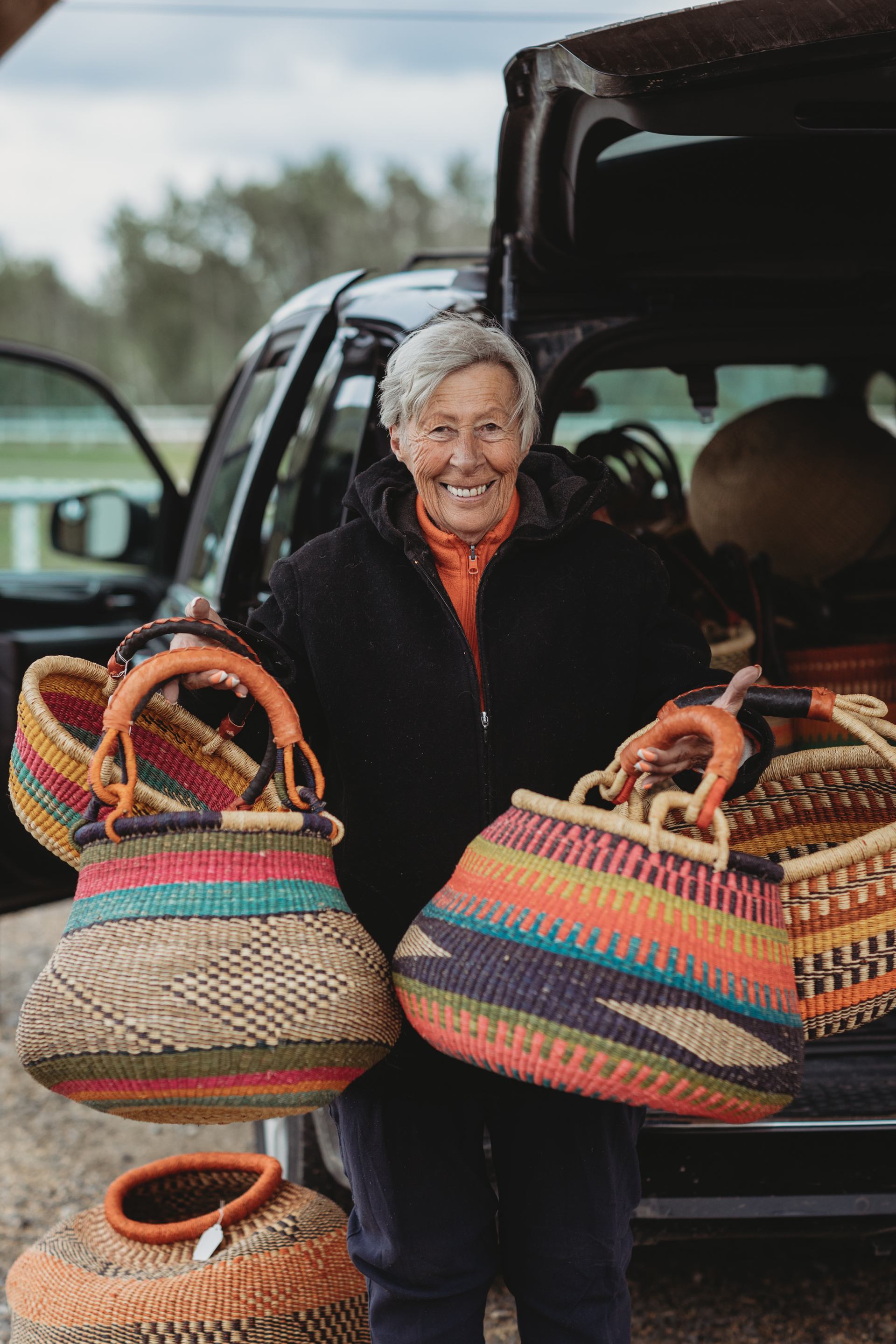 Person standing at the back of a vehicle holding two colourful handwoven baskets at an outdoor market.