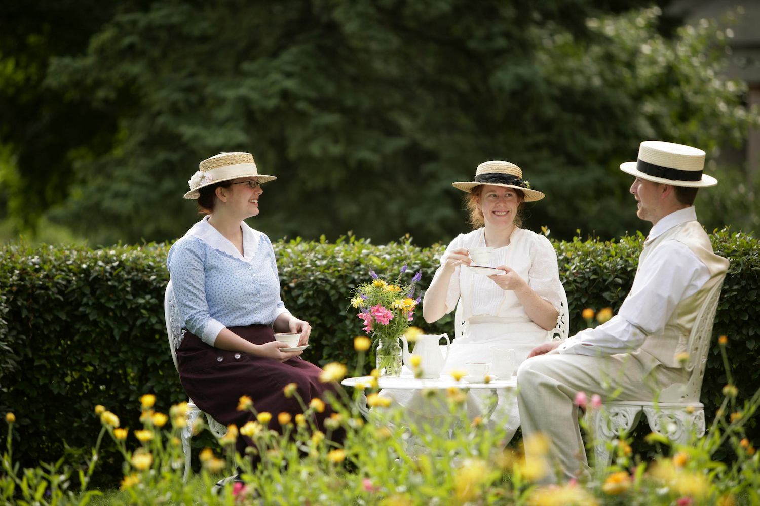 Three people in vintage clothing drink tea at a garden table with flowers.
