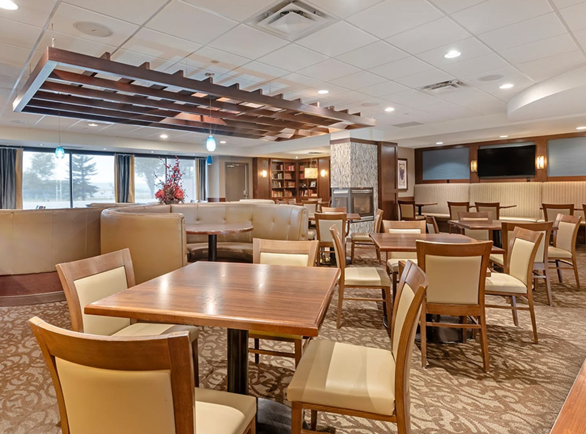 Hotel dining area with wooden tables, beige chairs, fireplace and bookshelf in background.