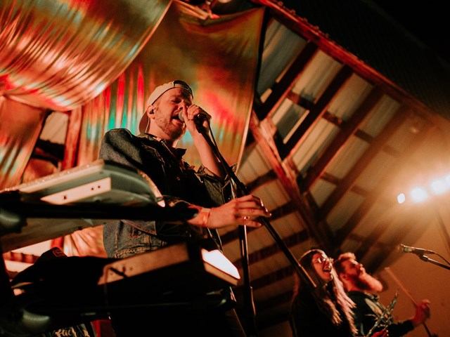Musician singing into a microphone on a stage with spotlights and instruments visible.